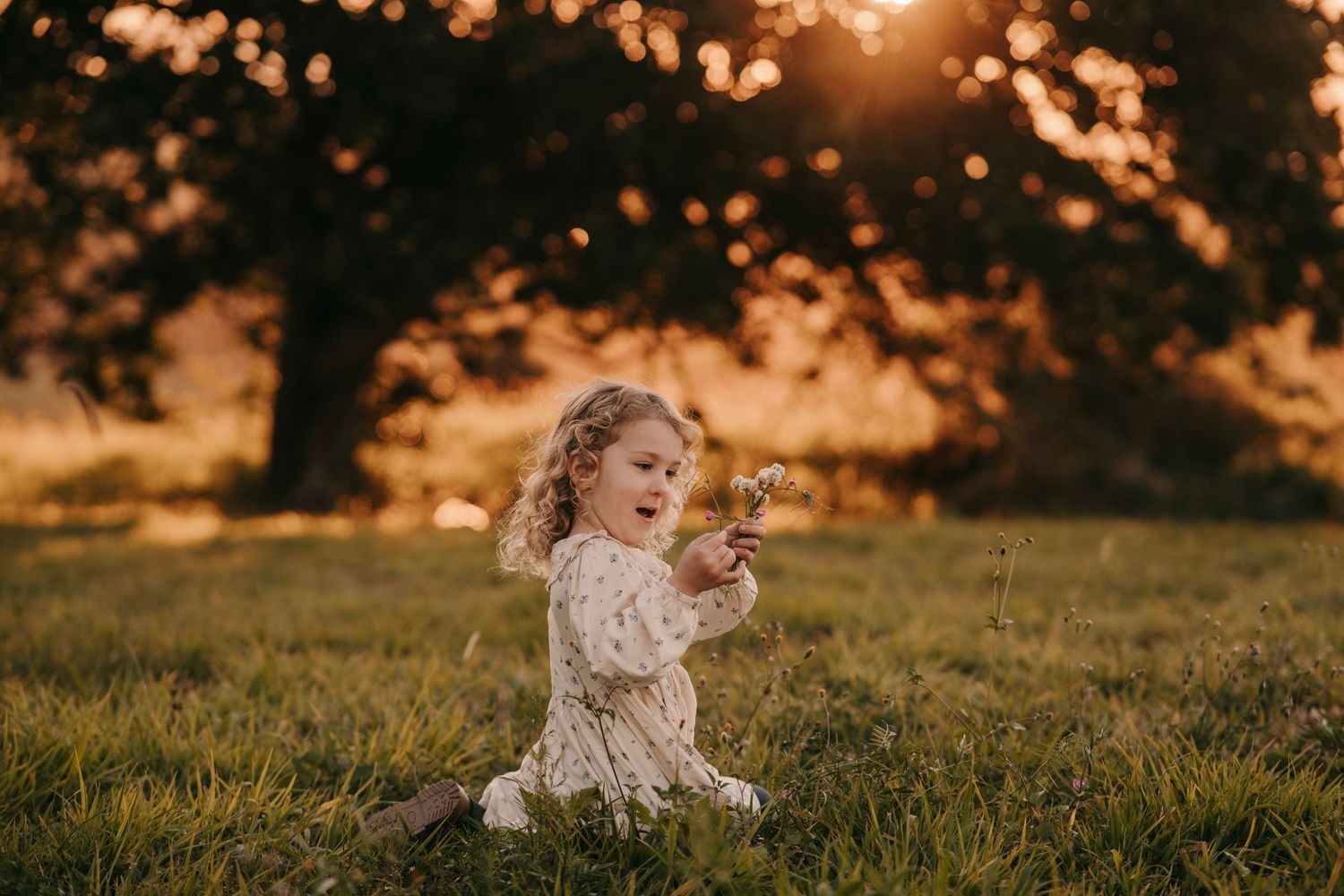 A young girl in a floral dress sits on green grass holding wildflowers during a golden sunset outdoors.