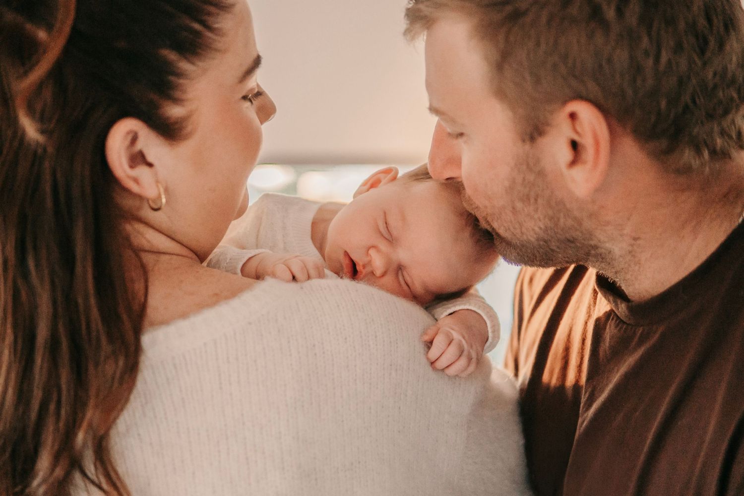 Loving parents leaning in to kiss their sleeping newborn baby in warm golden light.