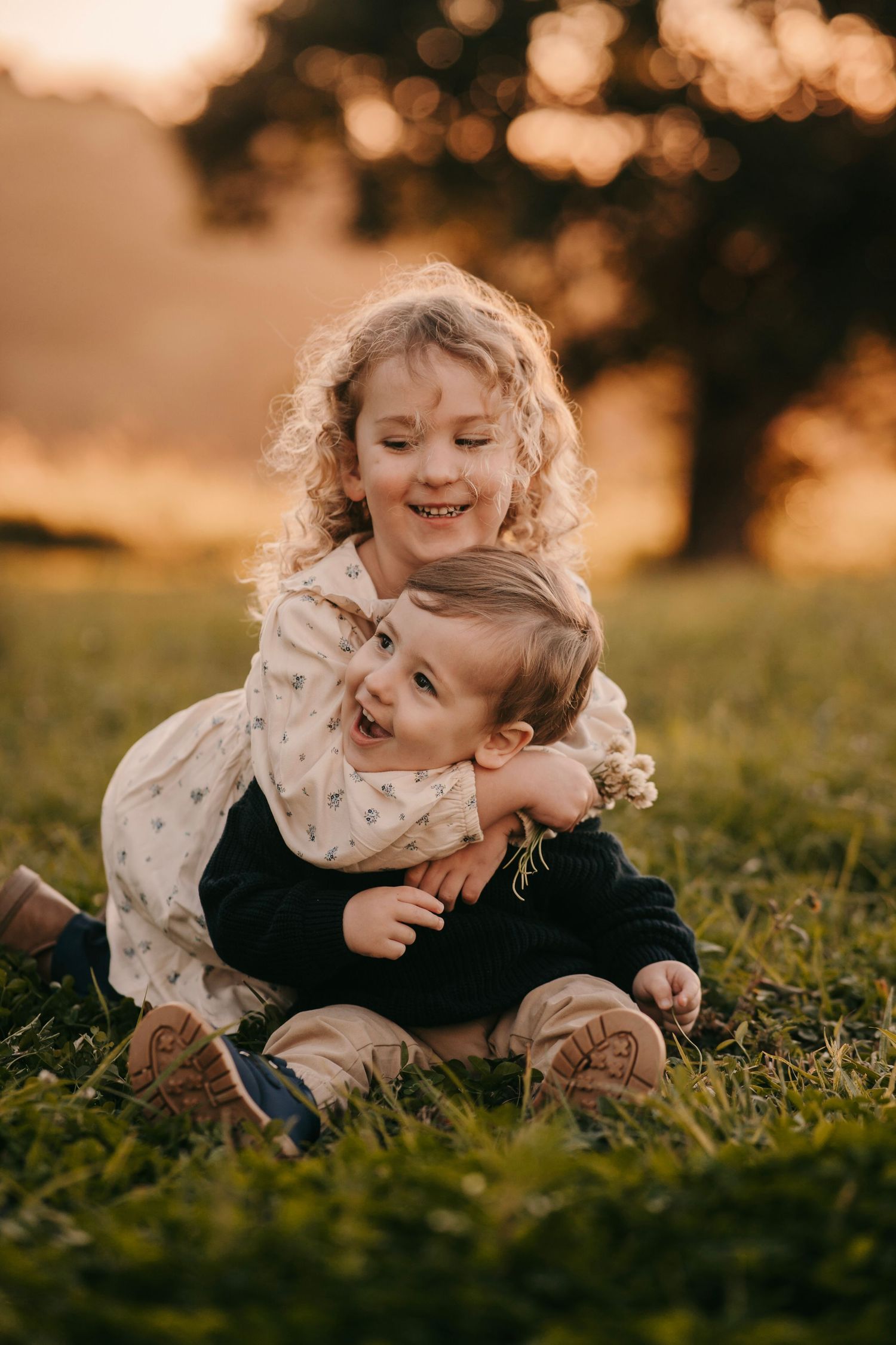 Two young siblings laugh and cuddle together on lush grass during a warm golden sunset.