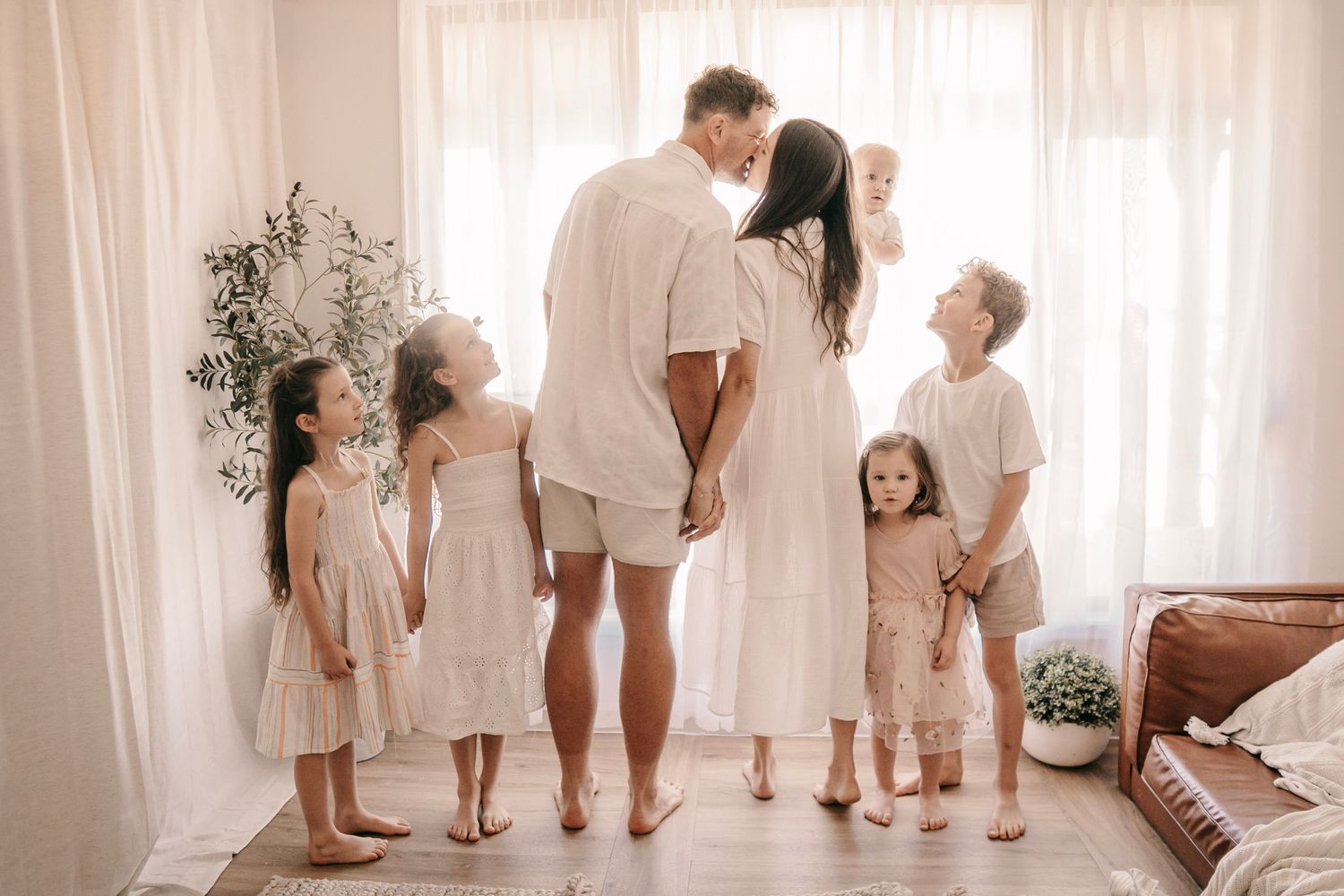 A family in white outfits shares a tender moment, with parents kissing surrounded by five children in a bright, airy room.