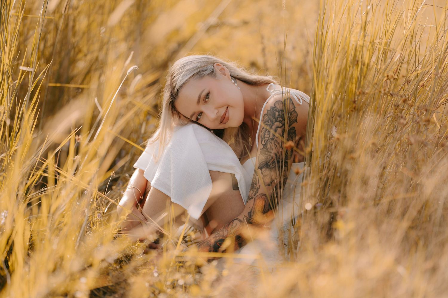 A smiling tattooed blonde woman sitting peacefully among tall golden grass in a white outfit.