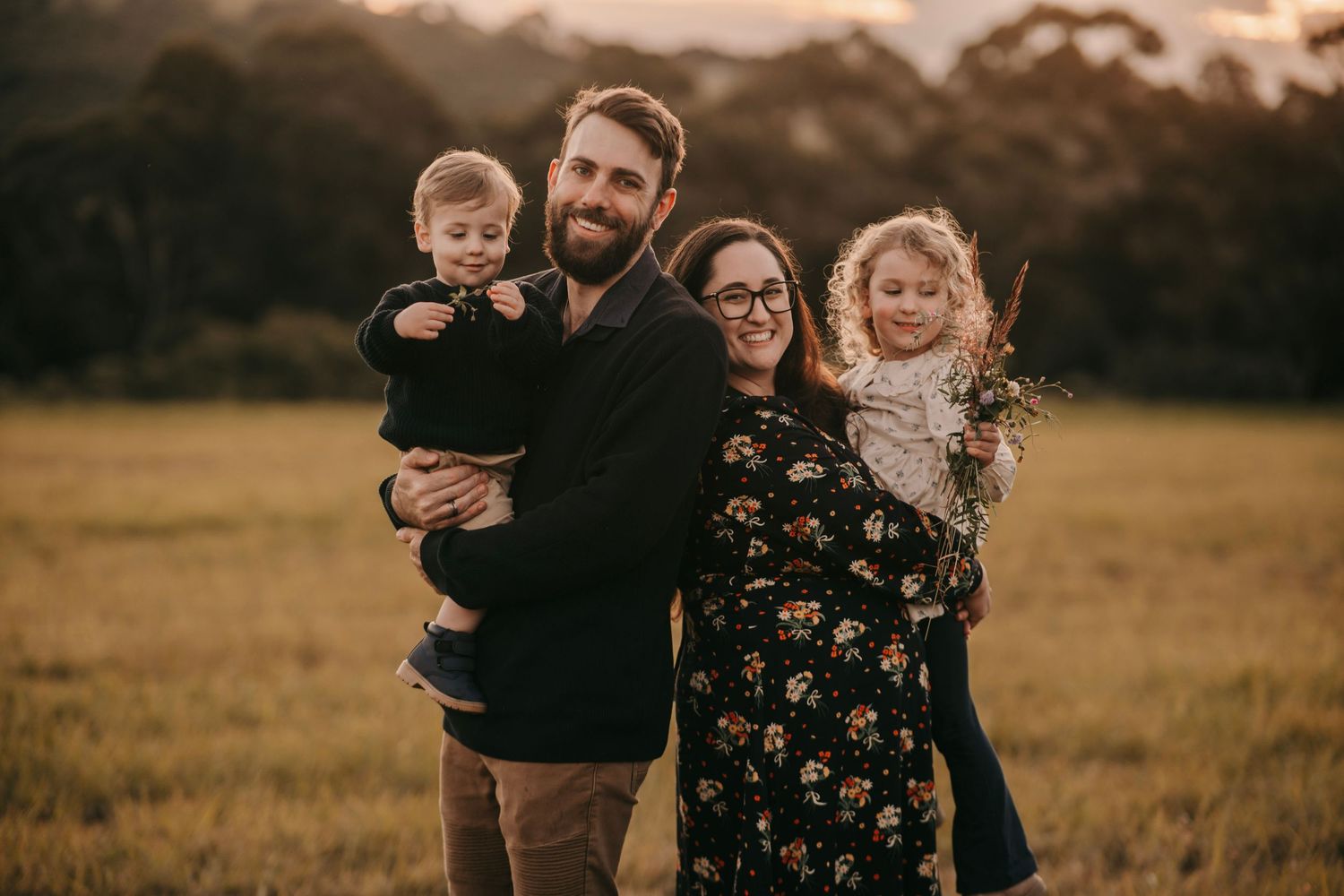 Smiling family of four posing together in an open field during golden hour.