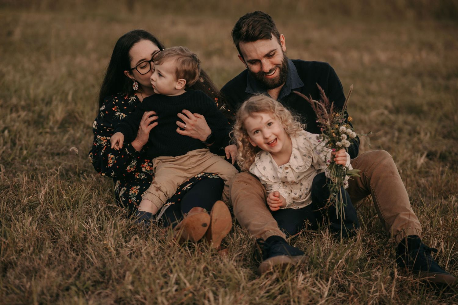 A happy family of four sits together in a grassy field during an autumn outdoor photo session.
