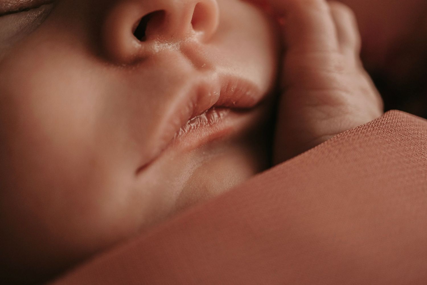 Close-up of a sleeping newborn baby's lips and tiny hand resting on a soft pink blanket.