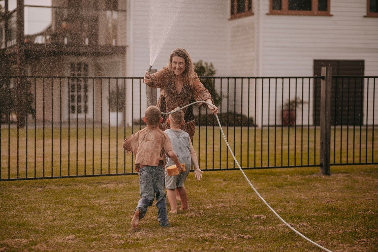 Joyful mother spraying children with a garden hose in a grassy backyard on a fun day.