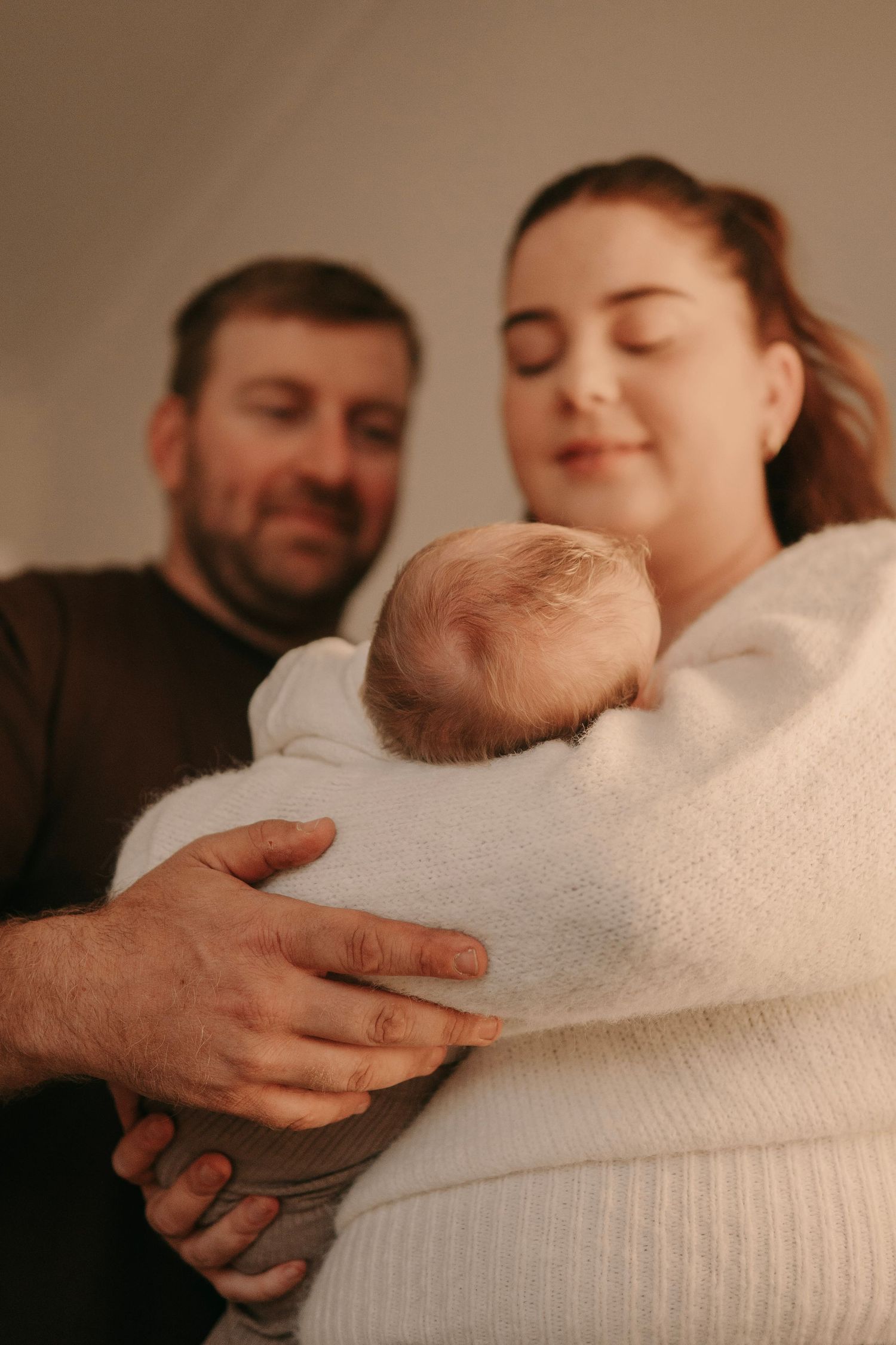 Parents with closed eyes lovingly cradling their newborn baby wrapped in a white knit blanket.