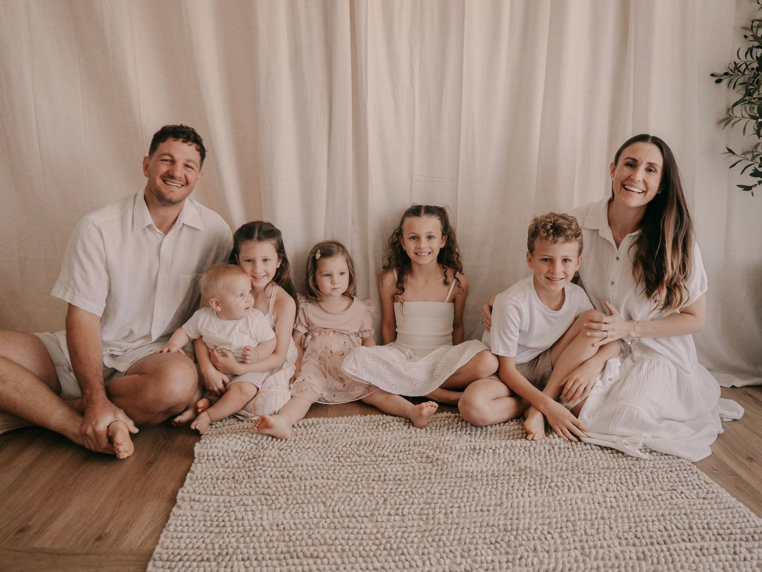Happy family of seven posing together on a rug against a white curtain backdrop in soft neutral tones.