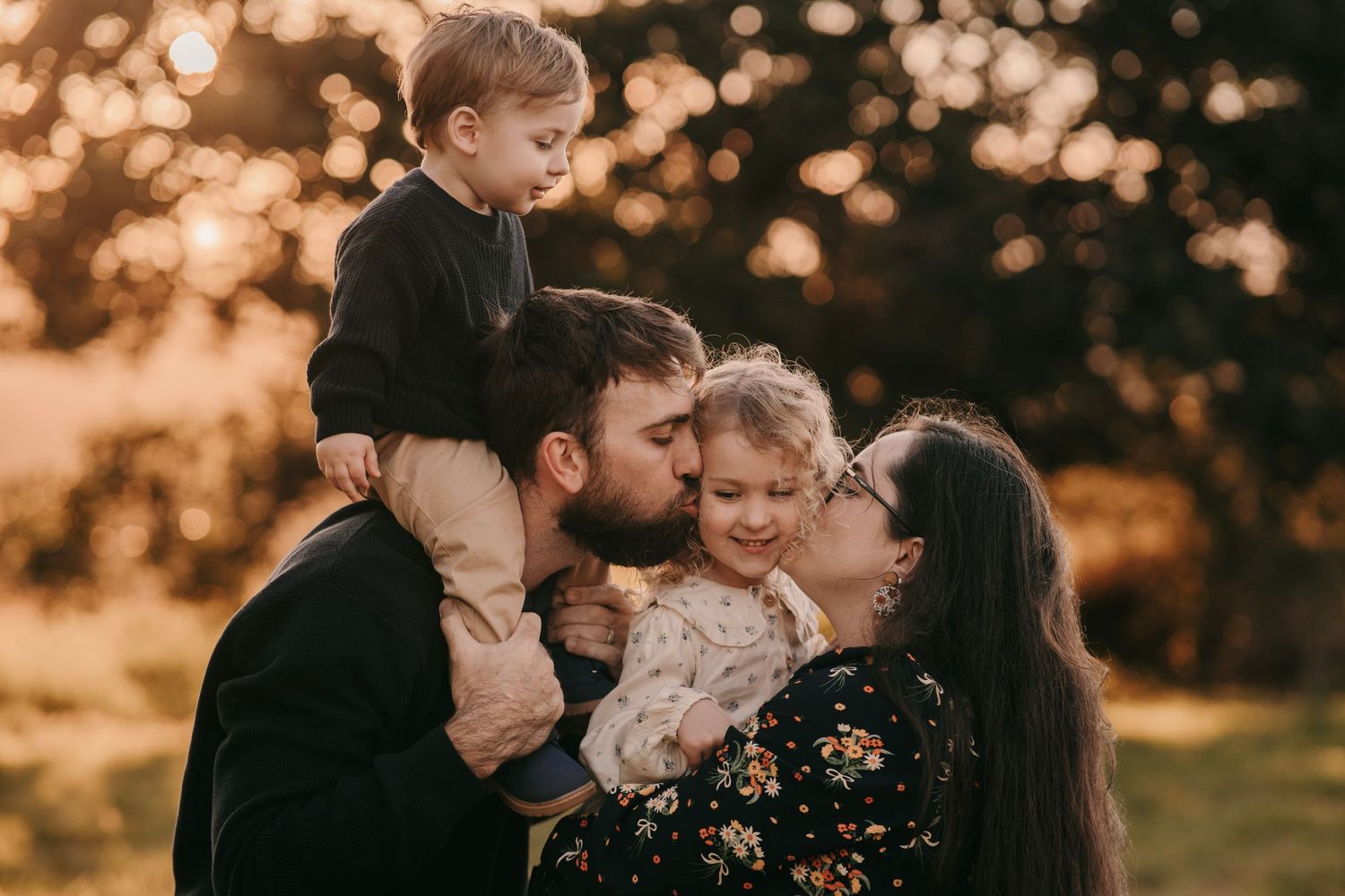 Family of four sharing kisses at golden hour in a park, parents holding young children in a warm embrace.