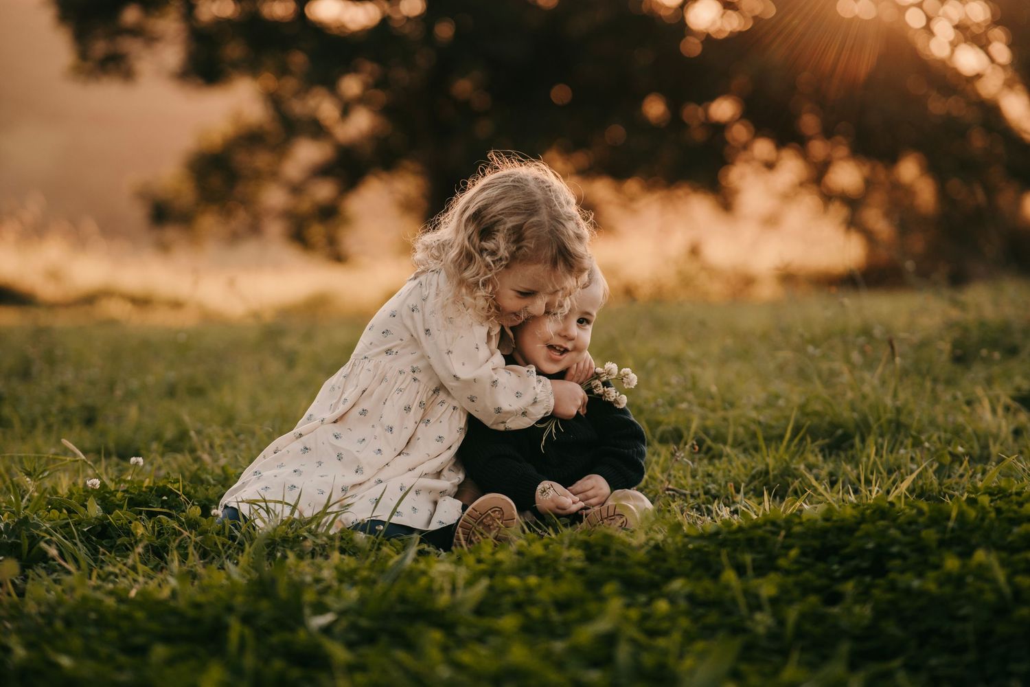 A curly-haired girl hugs a younger toddler sibling on green grass at golden hour sunset.