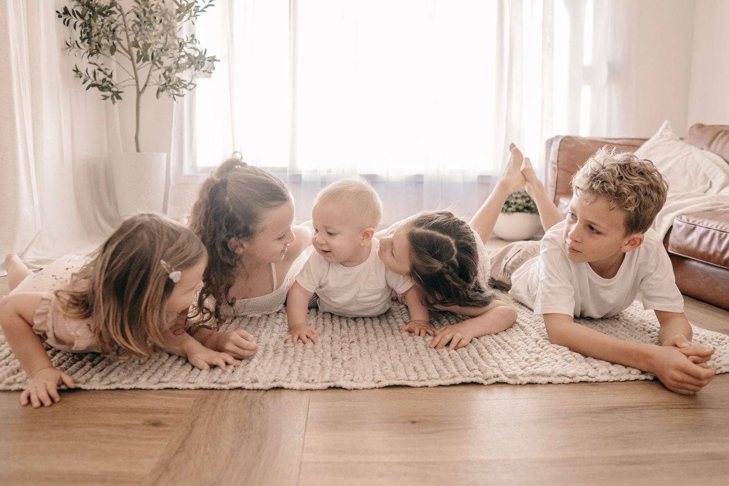 Four children lying on a rug, smiling and playing together in a bright, cozy living room.
