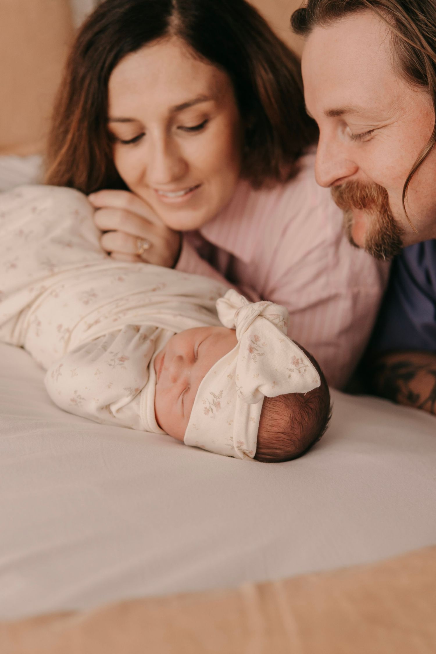 Smiling parents lying beside their swaddled newborn baby wrapped in floral blanket.