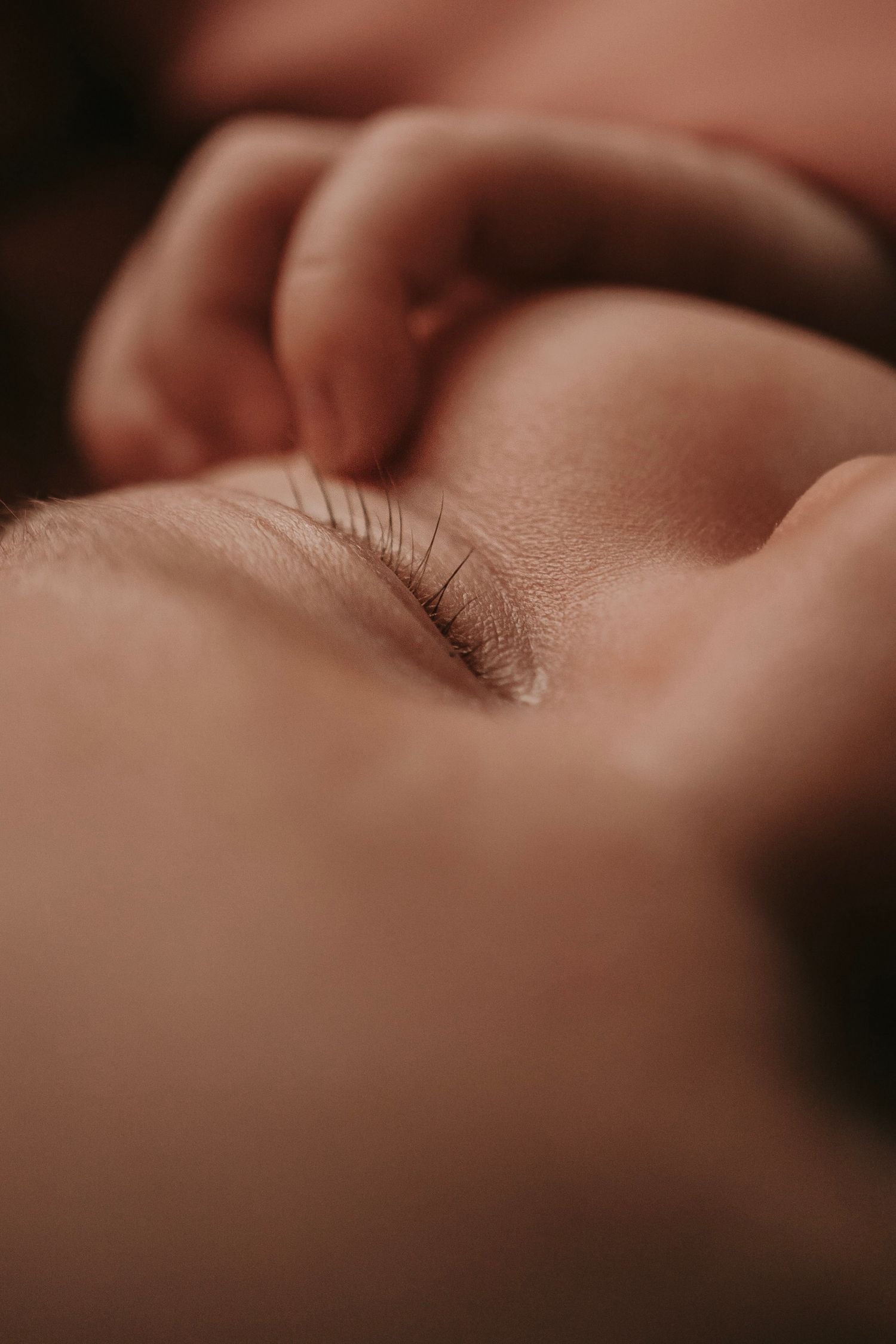 Close-up macro photo of a sleeping newborn baby's delicate eyelashes and tiny hand in soft warm light.