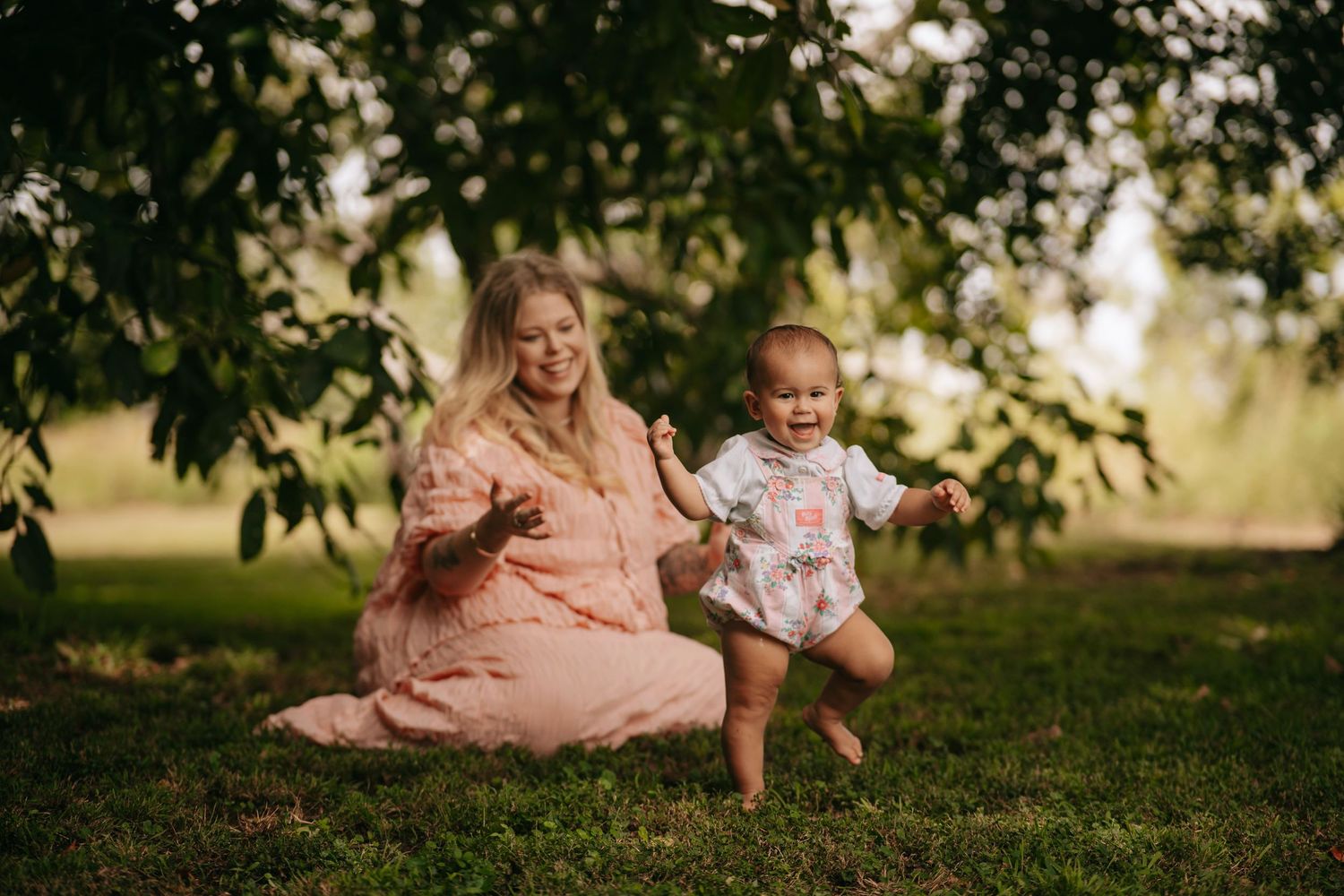 A blonde mother in a pink dress playing with her laughing toddler under a tree.