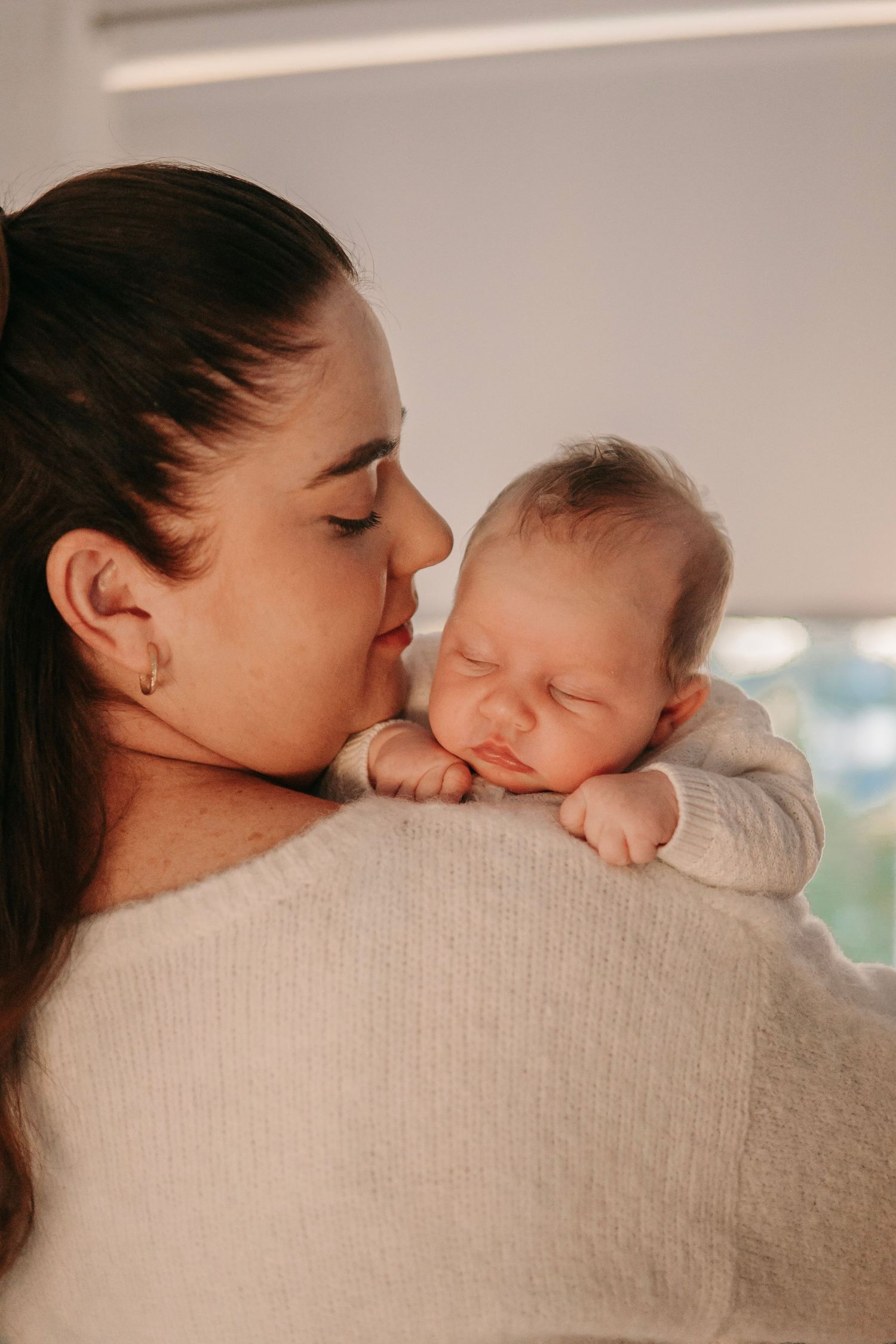 Mother tenderly holding sleeping newborn baby on her shoulder in soft natural light.