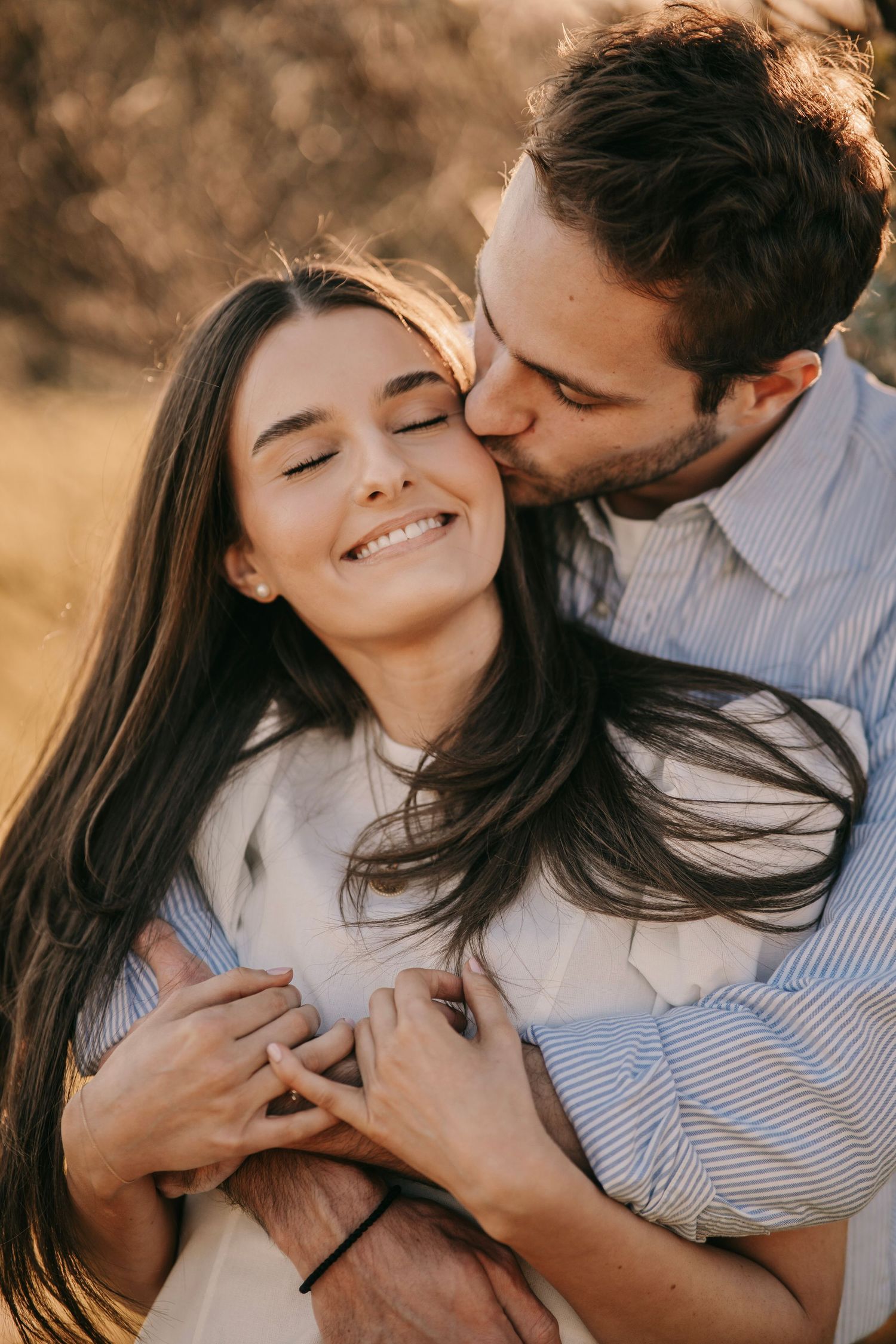 A man tenderly kisses a smiling woman on the cheek while embracing her from behind outdoors.
