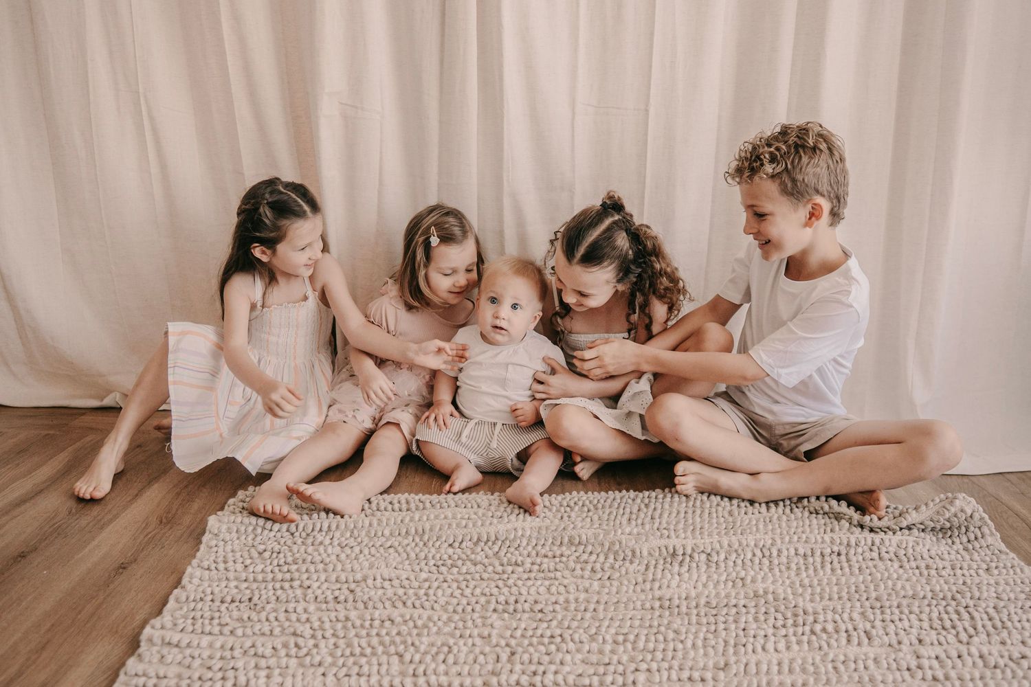 Five young siblings sitting together on a rug, lovingly interacting with their baby brother in a bright room.