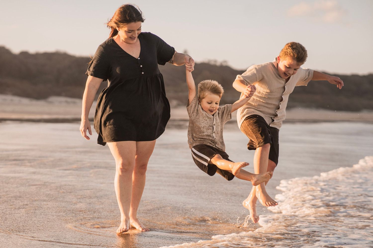 Mother swinging two children by the hands at the beach shoreline at sunset.