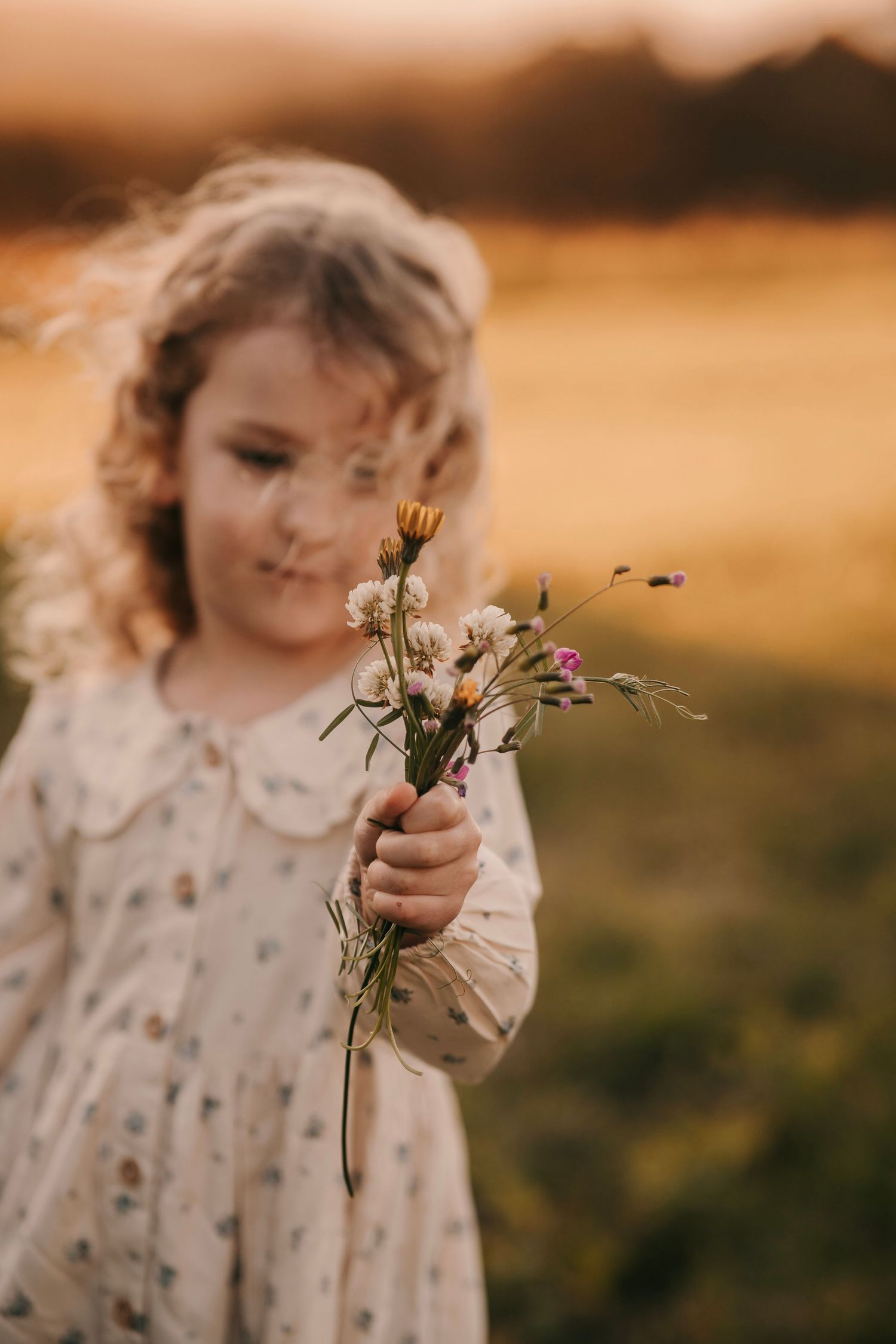 Young girl in floral dress holding wildflowers in a golden sunset field.