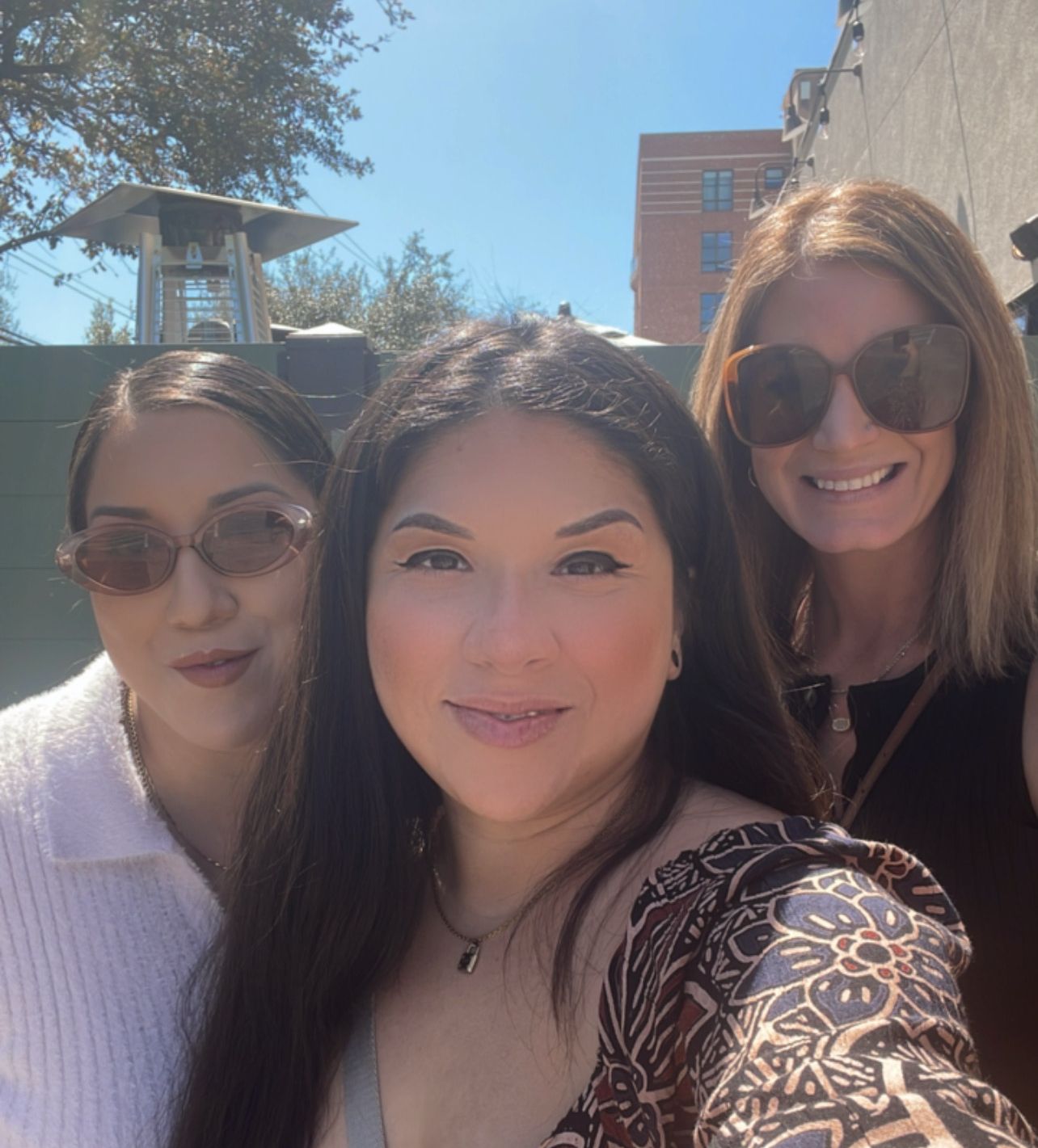 Three women smiling outdoors on a sunny day, capturing a casual group selfie