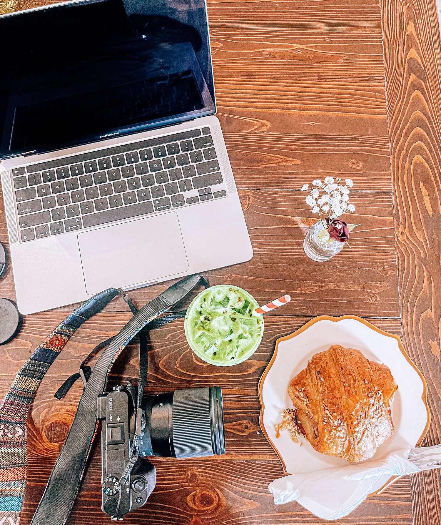 Flat lay of camera gear, iced matcha, pastry, and laptop on wooden café table for lifestyle content creation