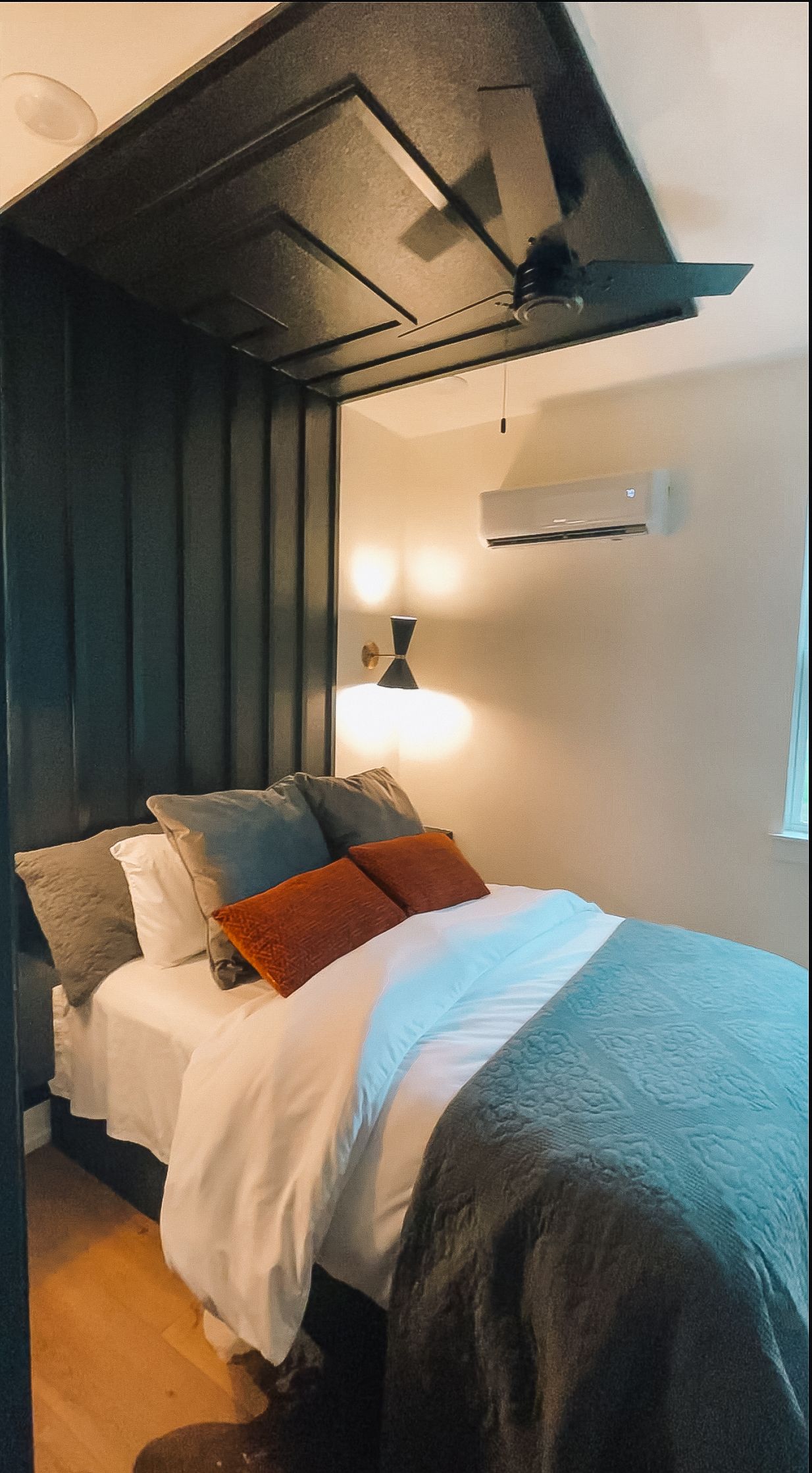 Modern bedroom with geometric ceiling design features blue and orange accent pillows on white bedding and a wall-mounted AC unit.