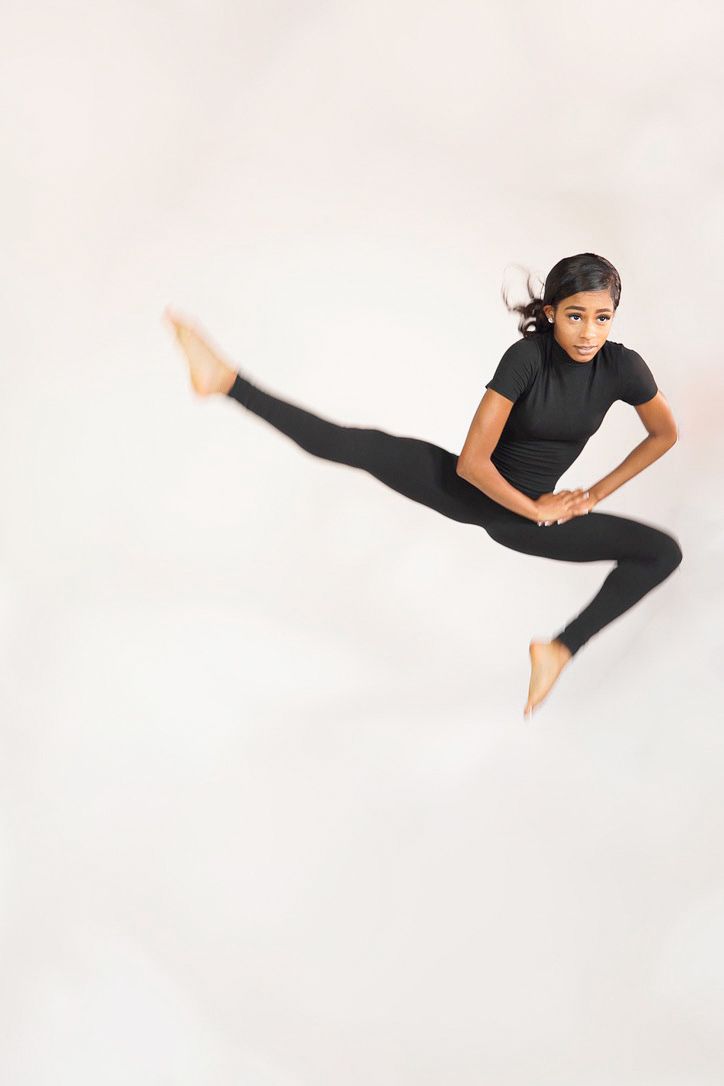 Young woman in black activewear captured mid-leap against a neutral backdrop, showcasing motion and confidence