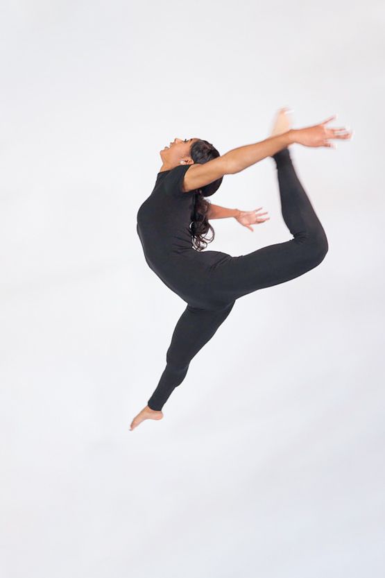 Dancer in mid-air leap with bent leg and arms lifted, wearing all black against a soft backdrop