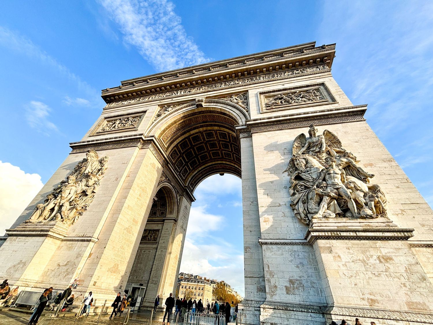 Wide-angle view of the Arc de Triomphe under a vivid blue sky with passing tourists