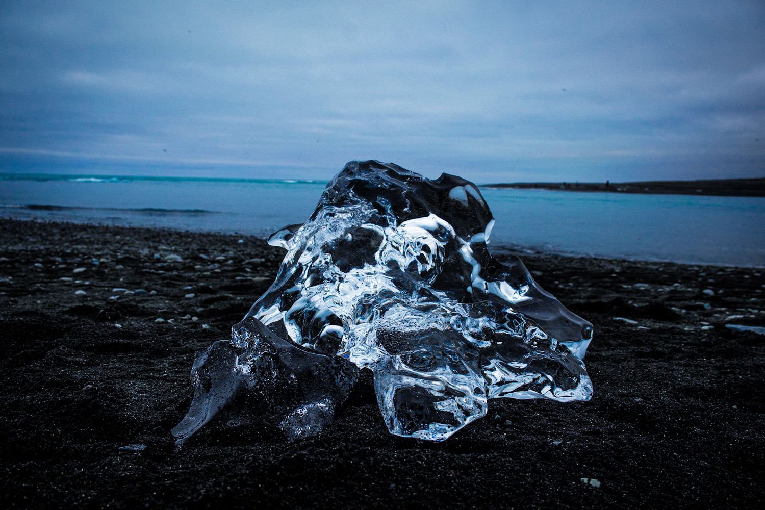 Chunk of clear ice resting on black sand beach under a moody blue sky