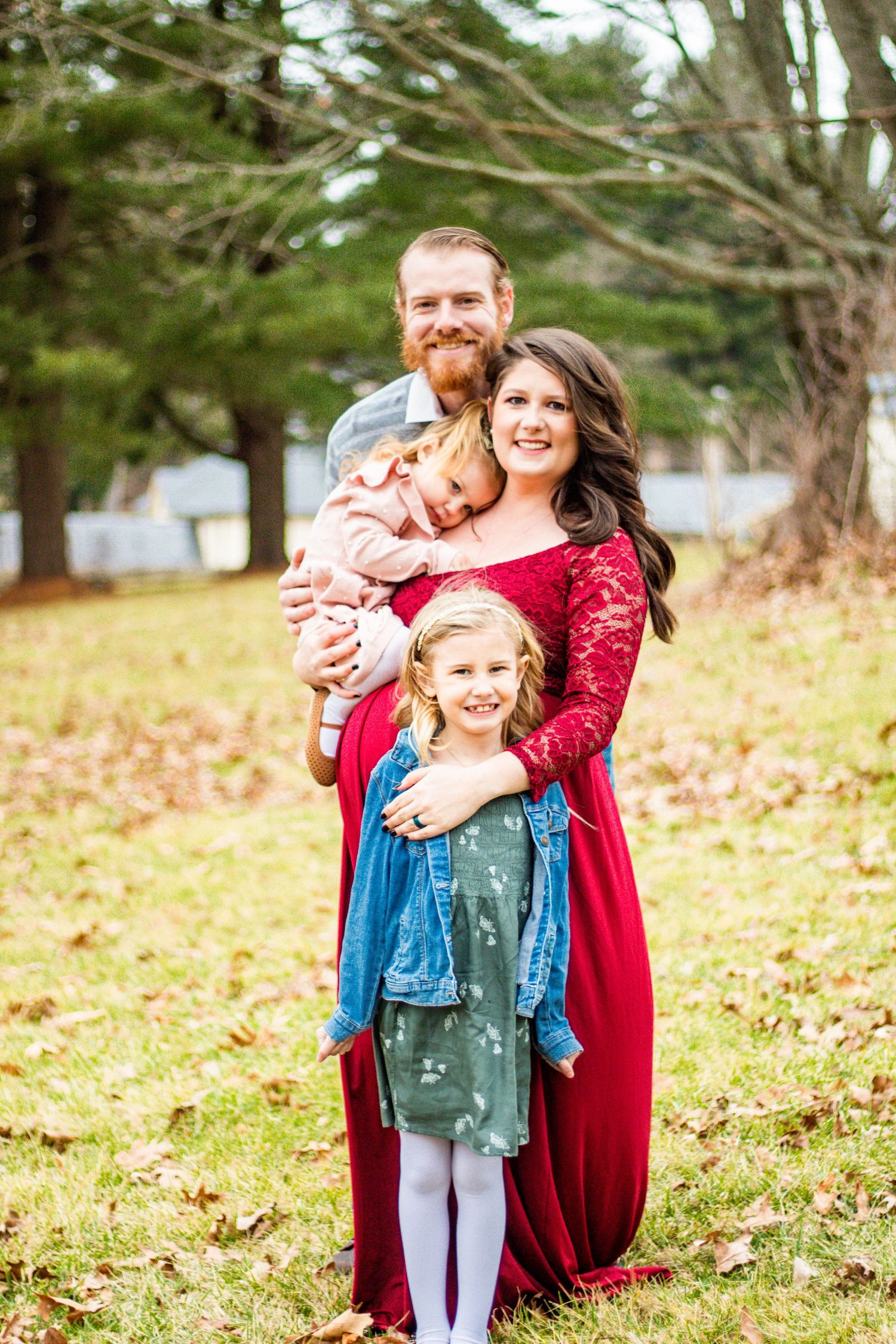 Family portrait outdoors in autumn with fallen leaves and evergreen trees in background.