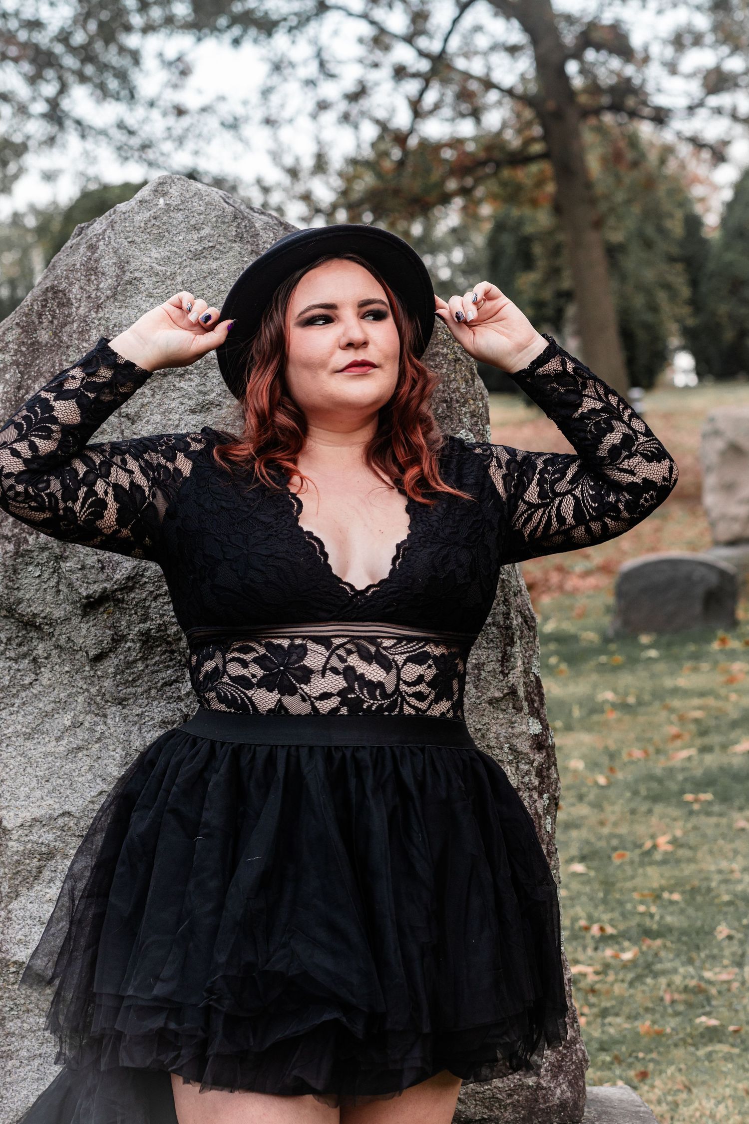 Gothic style black lace and tulle dress with hat photographed against large stone in outdoor cemetery setting.