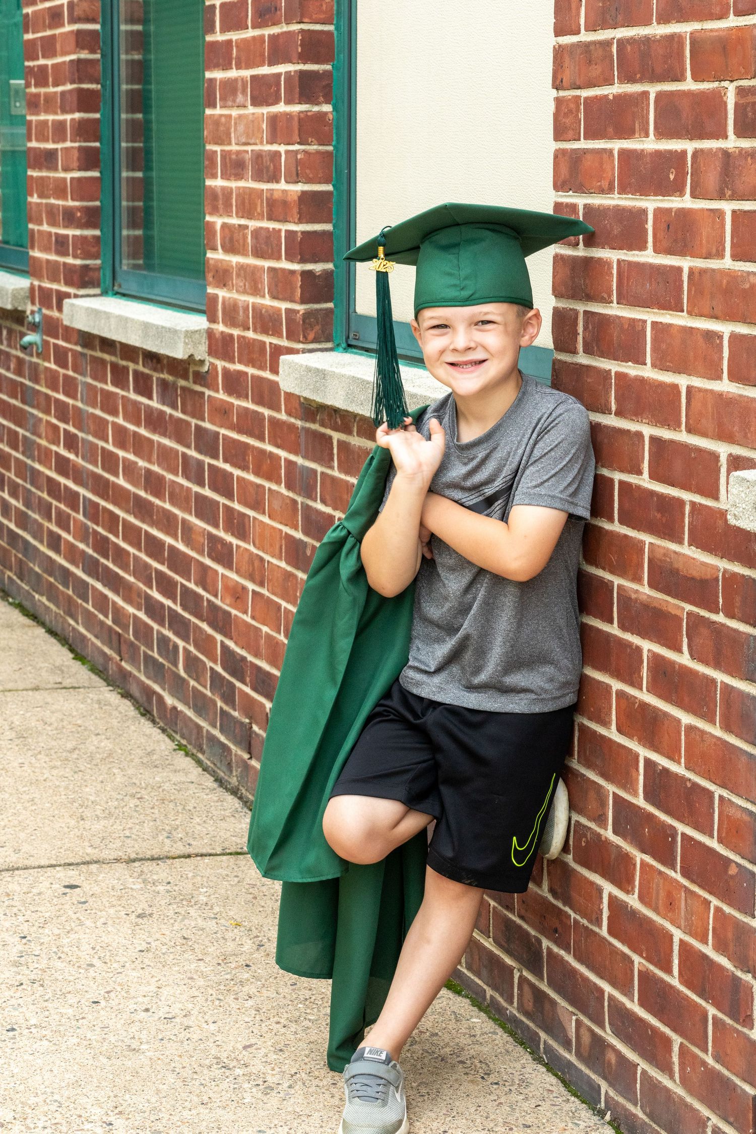 Child in gray shirt posing with green graduation cap and gown against brick wall.