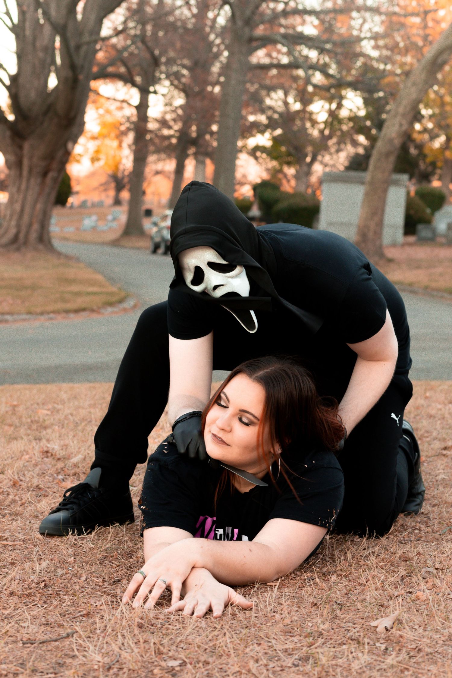 Horror themed photoshoot with person in ghostface costume and subject on autumn ground.