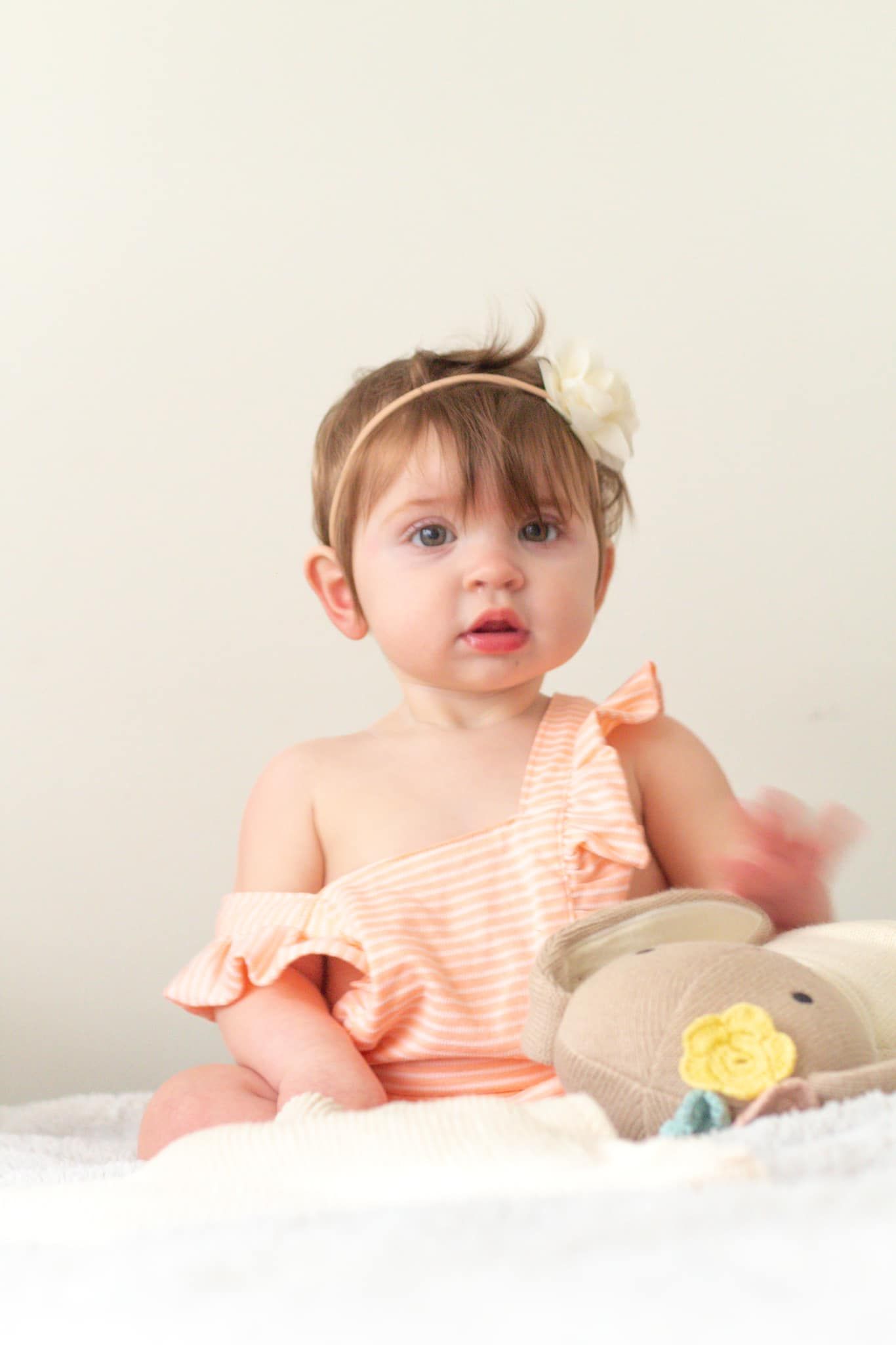 Baby in peach striped dress lying on white blanket with floral accessories.