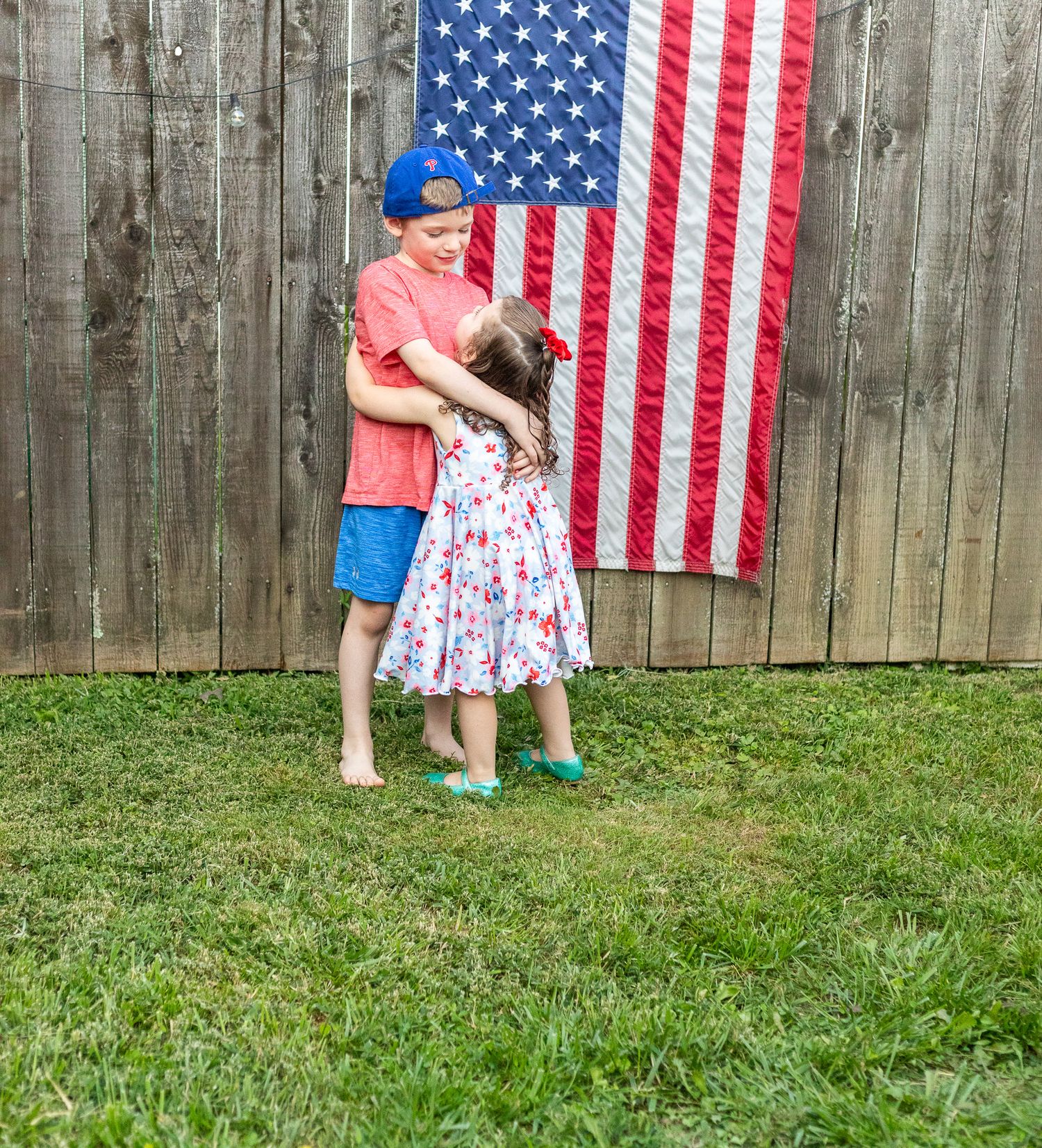 Two people dance together in front of a large American flag hanging on a wooden fence in a backyard.