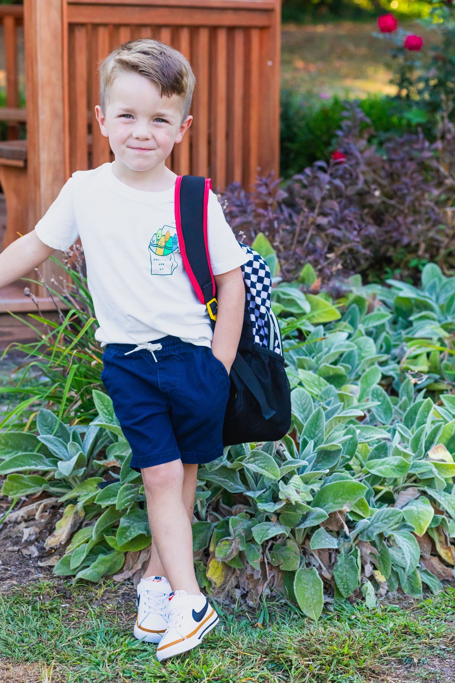 Young student with backpack poses in garden wearing navy shorts and white t-shirt.