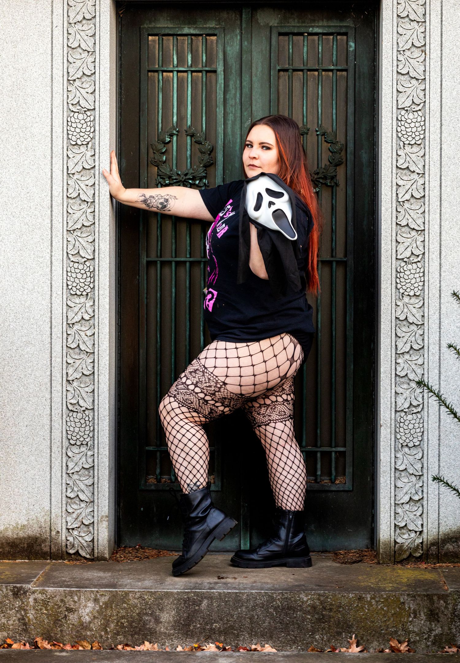 Figure in black outfit with fishnet stockings and gothic style boots poses dramatically in front of ornate green double doors.