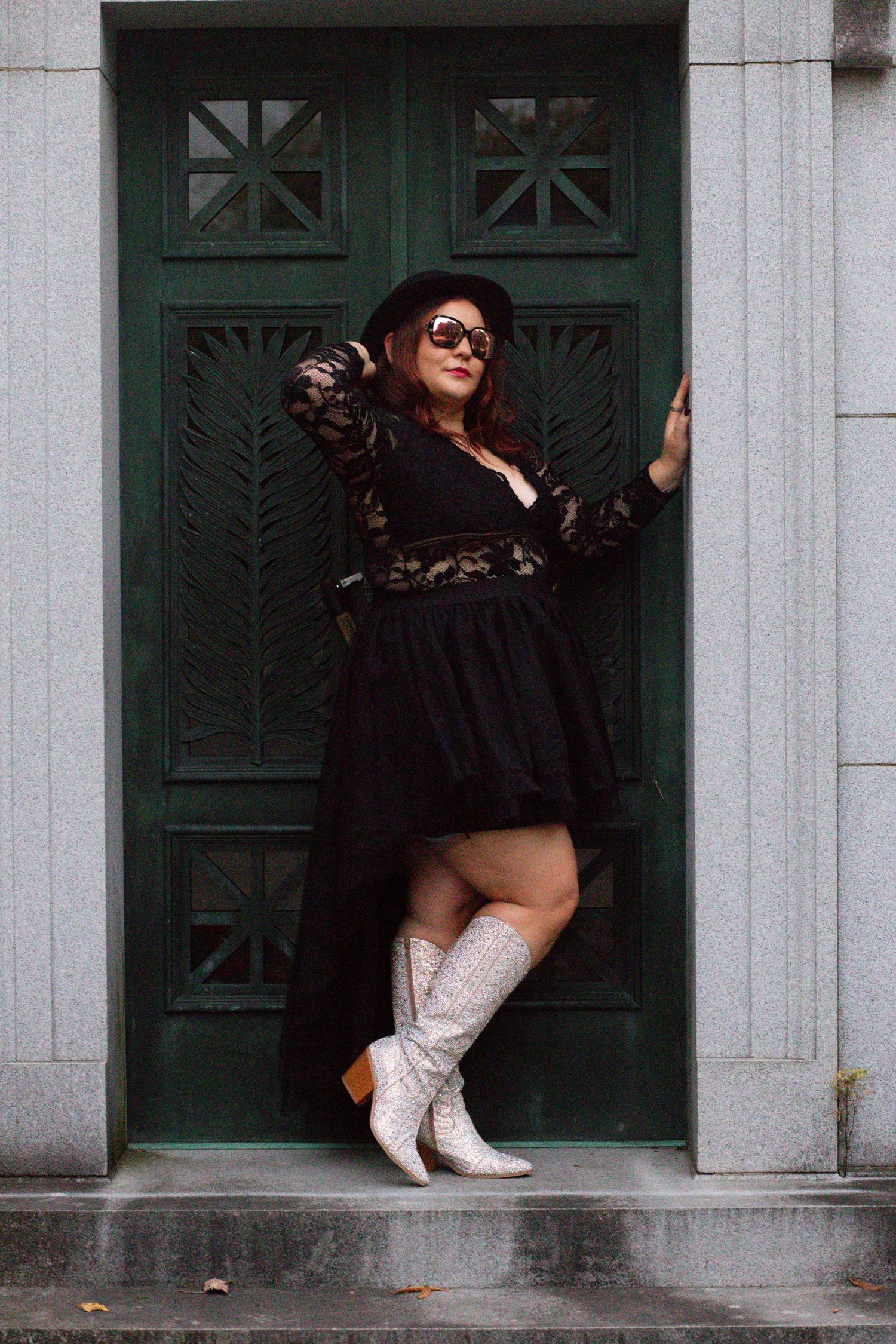 A person in a black dress and white cowboy boots poses against a decorative green door.