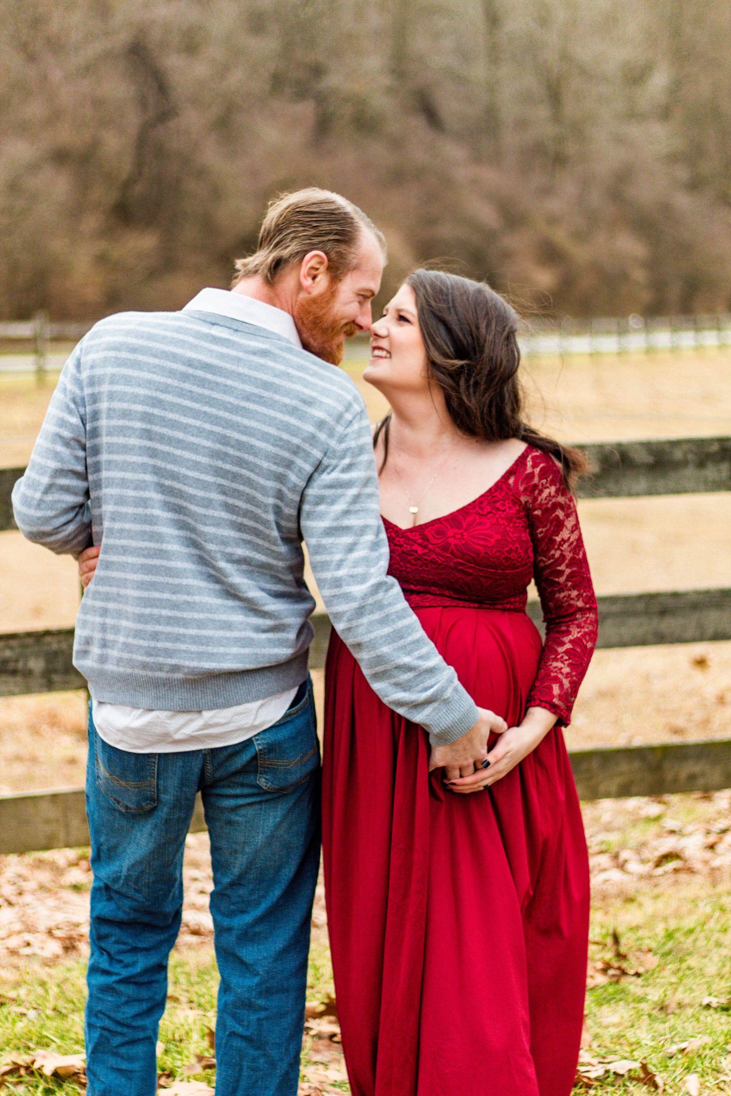 A romantic couple poses together outdoors, with her in a red lace dress and him in a striped shirt by a wooden fence.