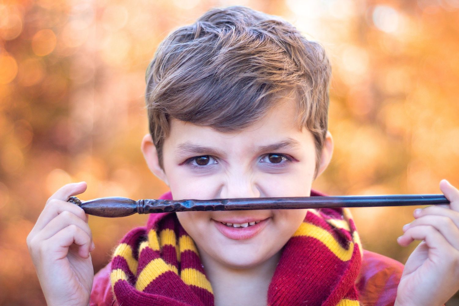 Child wearing striped scarf holds pencil across face with autumn bokeh background.