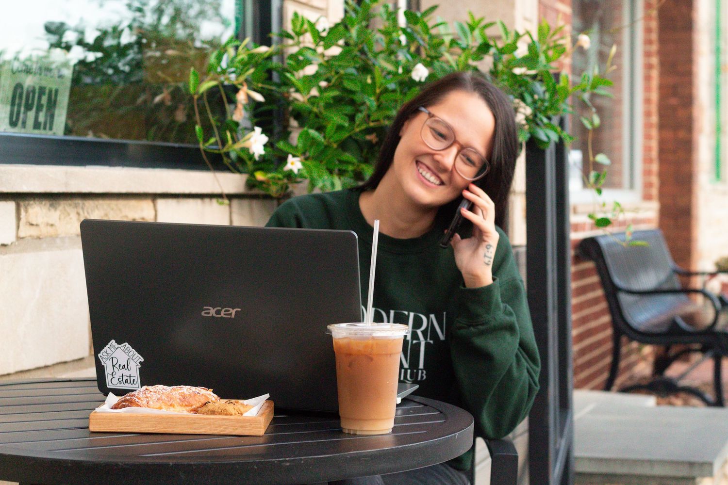 Someone in a green sweater works on a laptop at an outdoor cafe table with an iced coffee and sandwich.