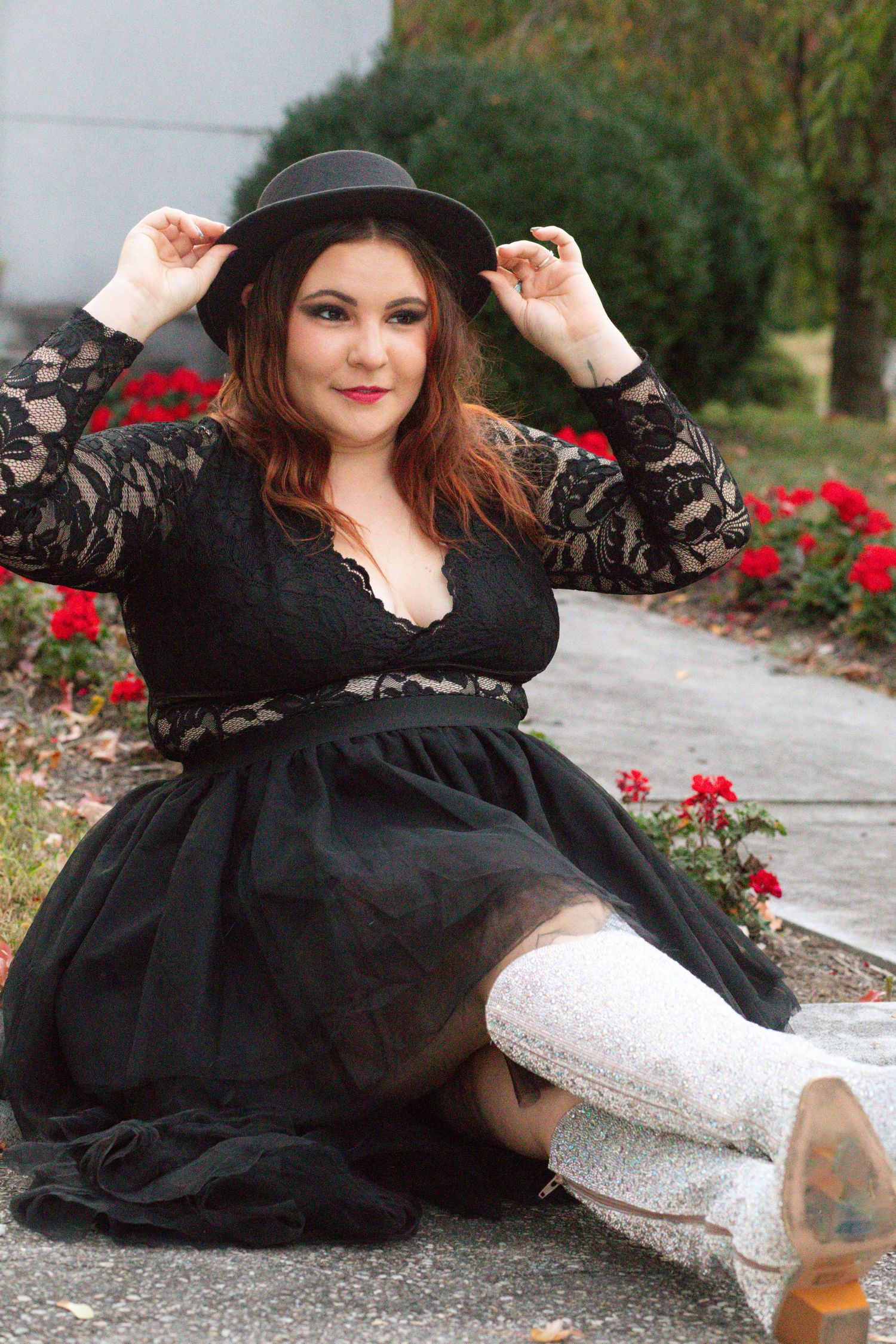 Person in black lace dress and wide-brimmed hat poses with white boots among red flowers in garden path.