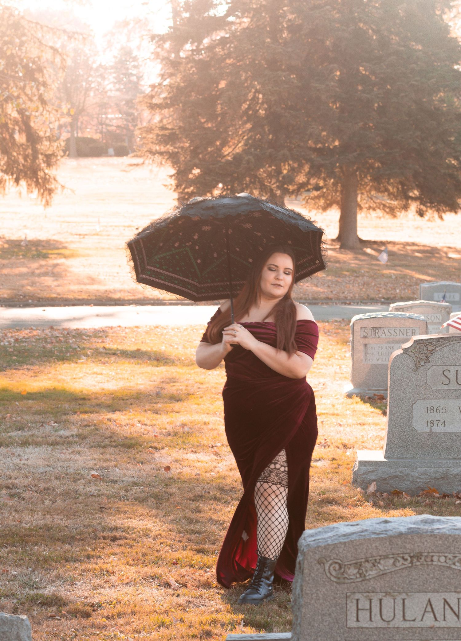 Person in velvet dress poses with umbrella in misty cemetery at sunset.