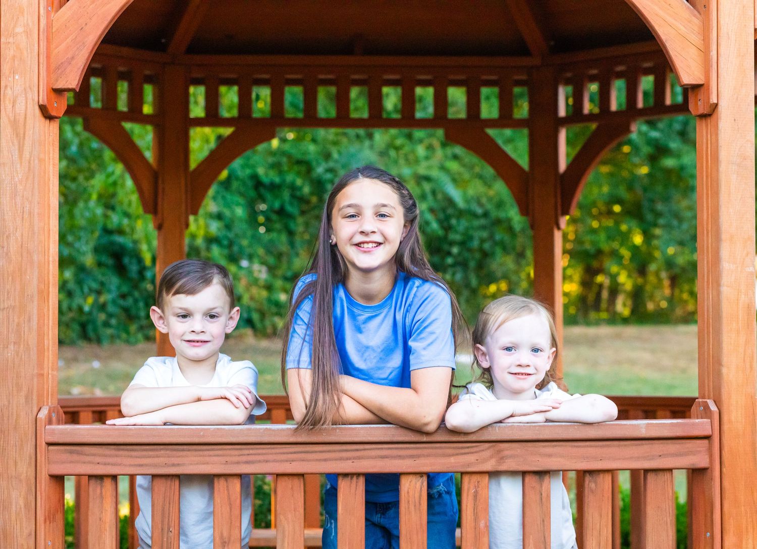 Group of people sitting on wooden porch benches at a park gazebo during a sunny summer day.