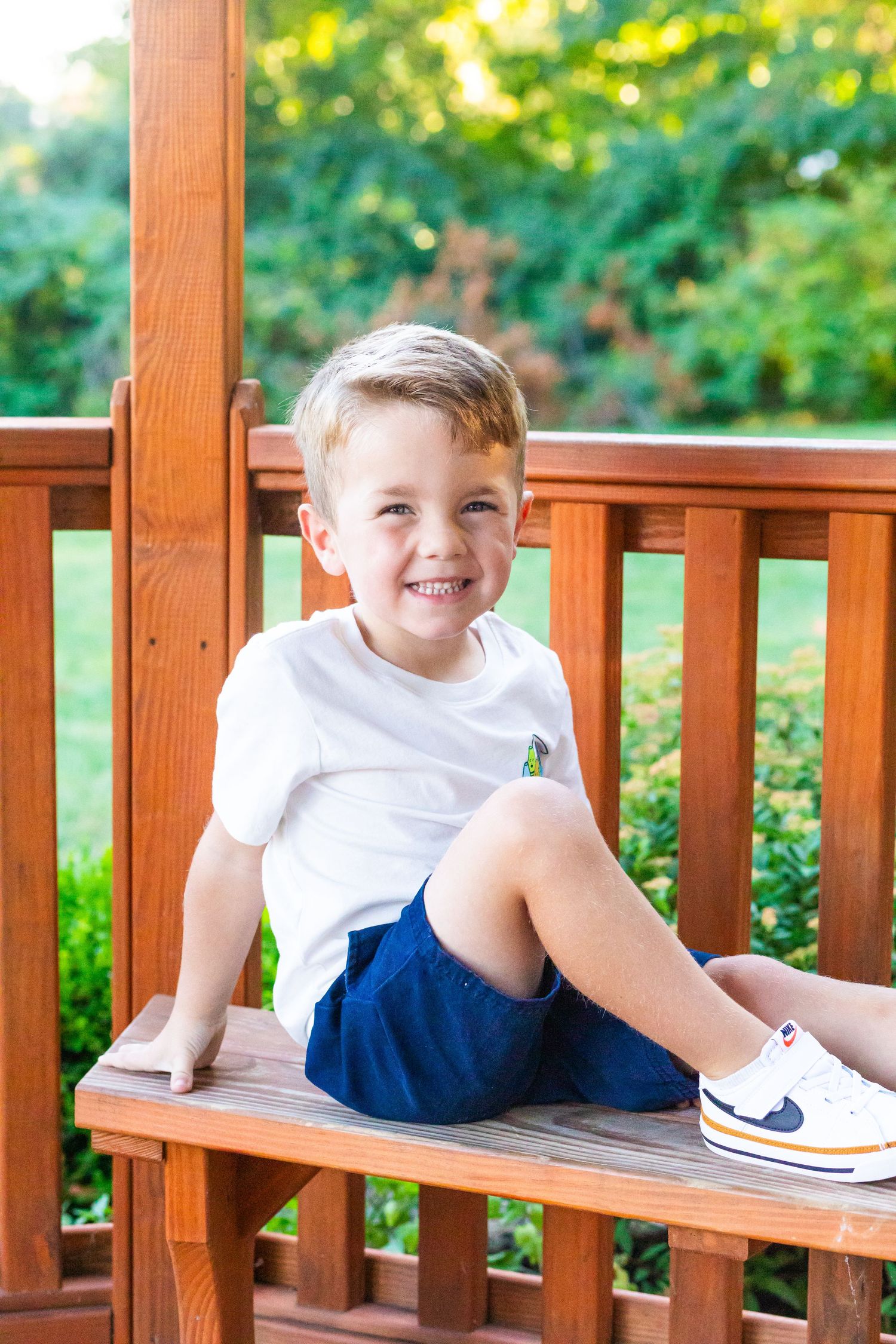 Young boy in white shirt and blue shorts sits casually on wooden bench outdoors with greenery in background.