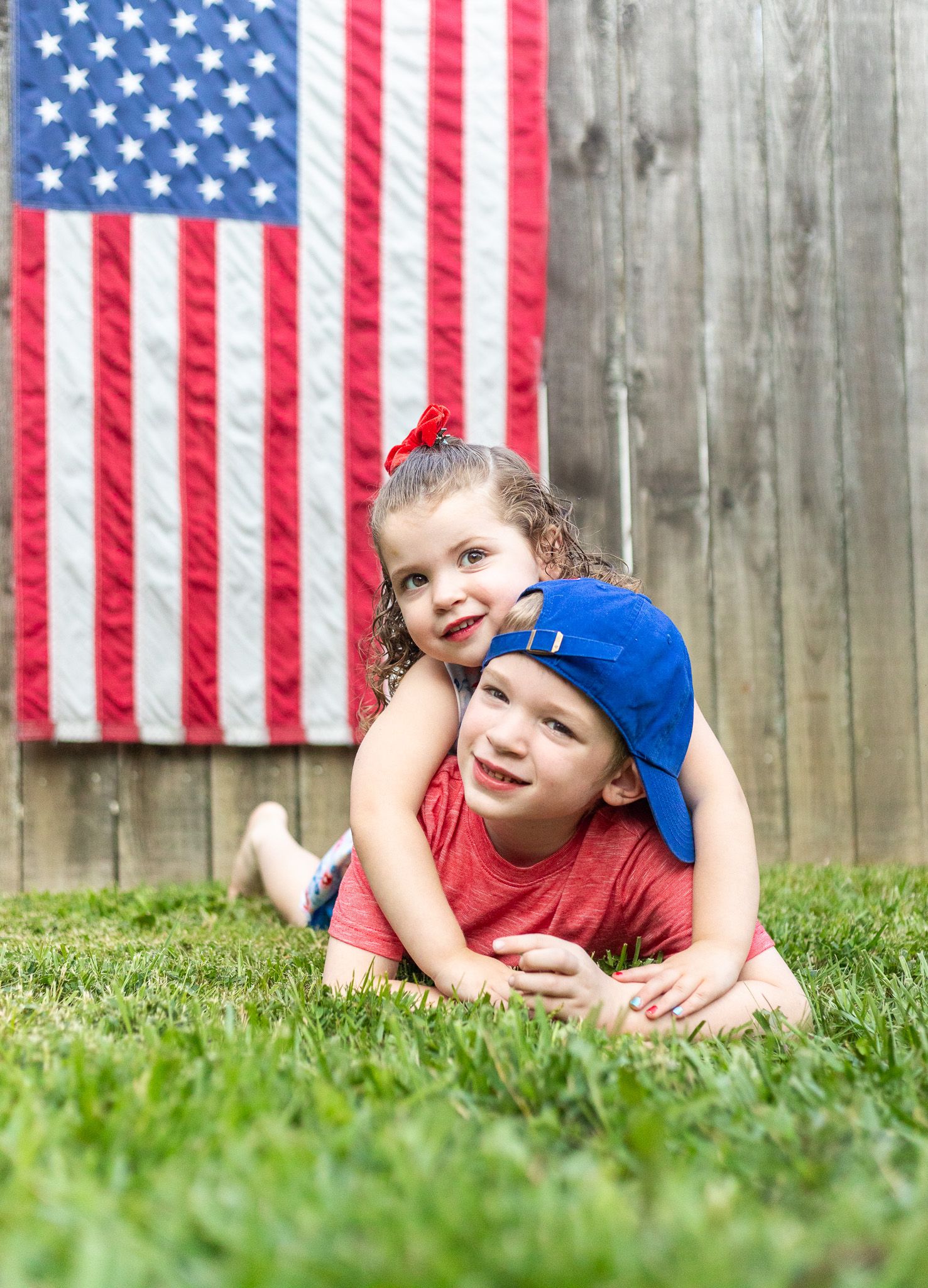 Two children playing on grass in front of American flag on wooden fence.