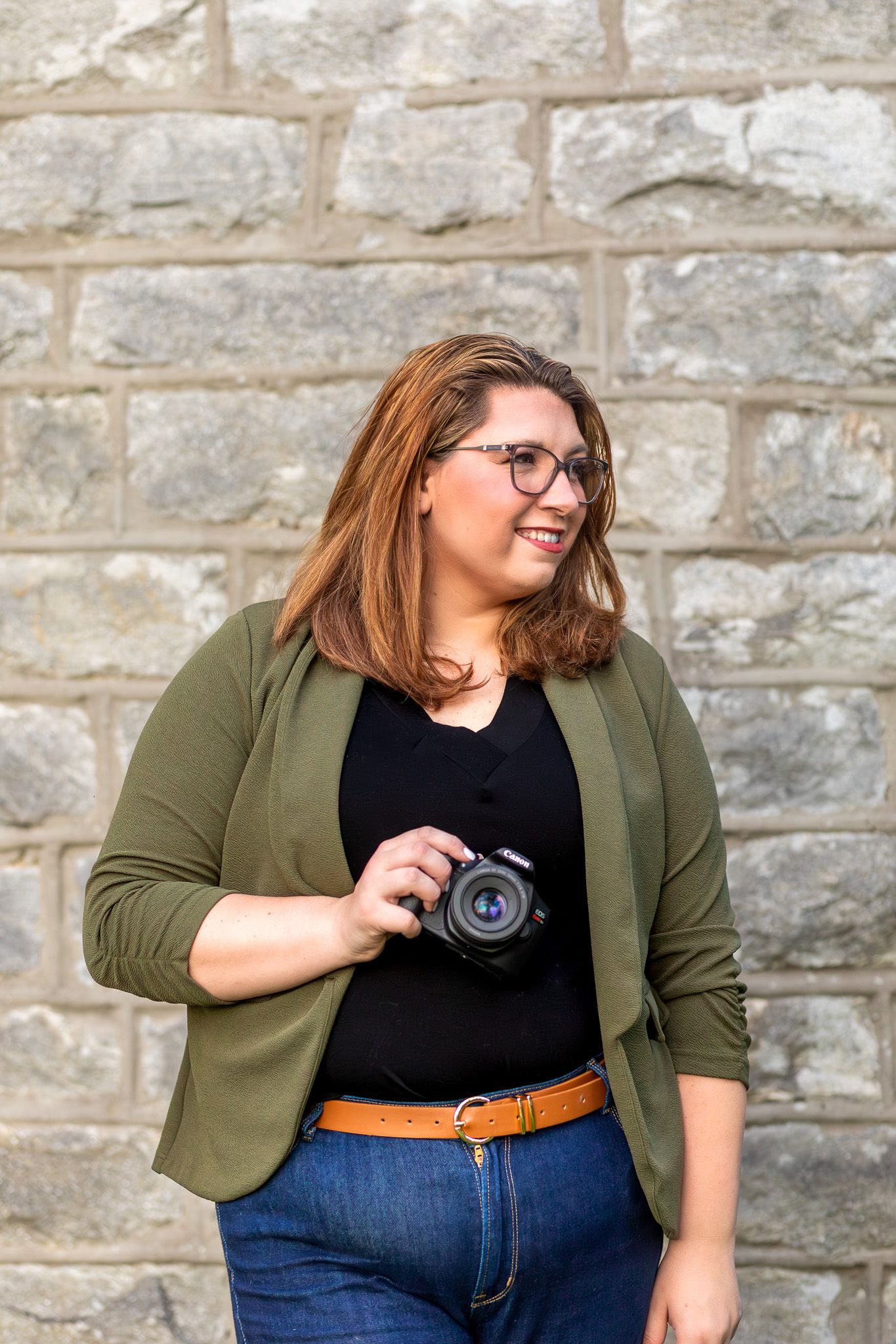 Photographer in olive blazer and jeans holding camera against stone wall.