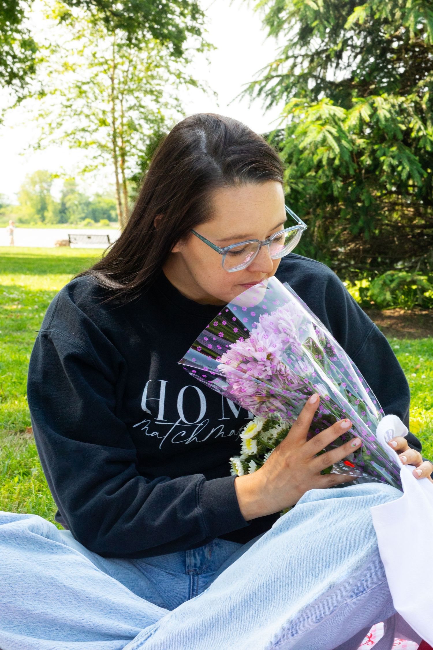 Someone smelling a bouquet of pink flowers while sitting on grass wearing a black sweatshirt.