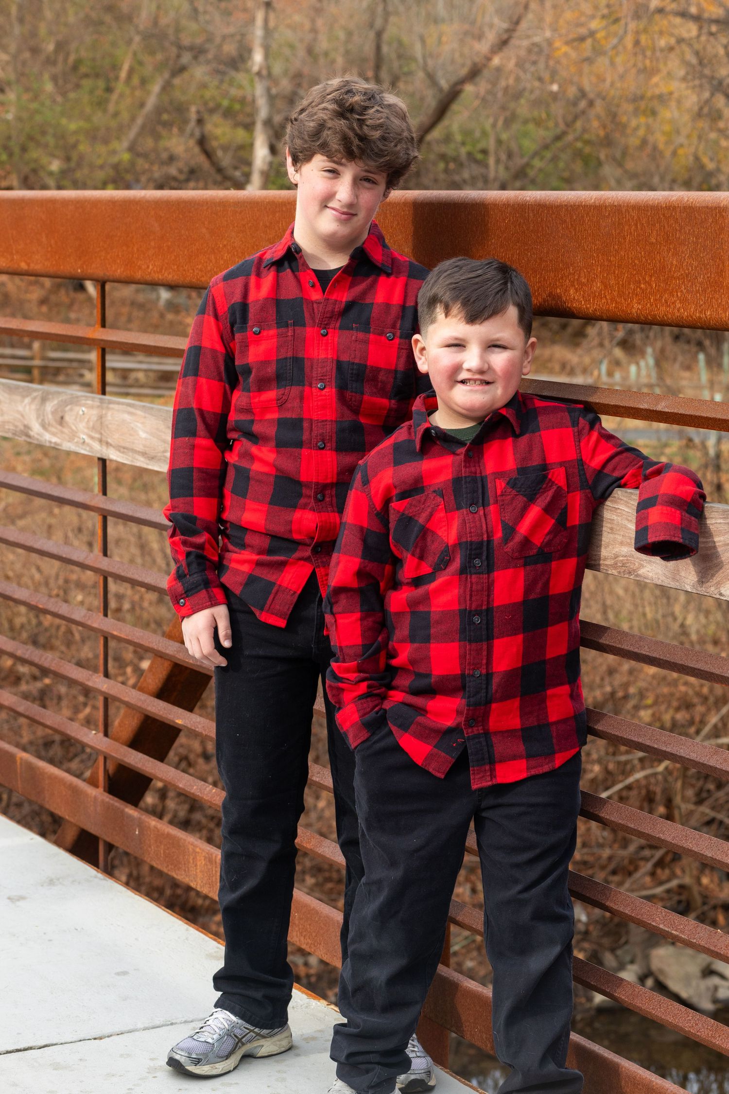 Two children in matching red buffalo plaid shirts and dark pants pose on a wooden bridge in autumn.