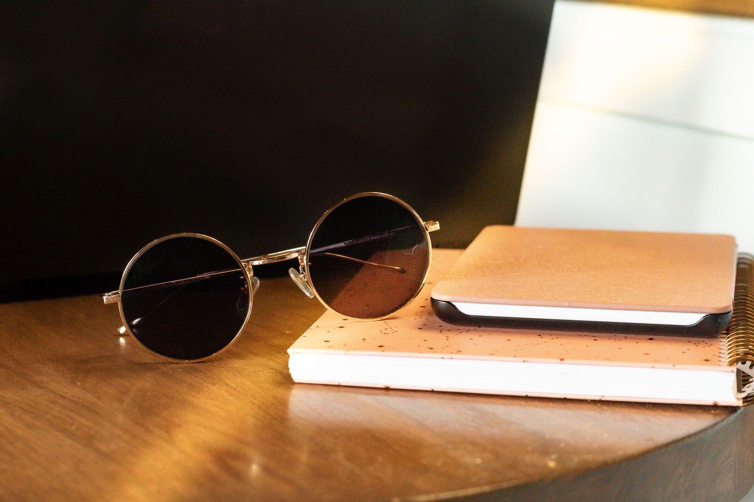 Round vintage sunglasses with brown lenses rest on wooden table next to leather notebooks in warm lighting.